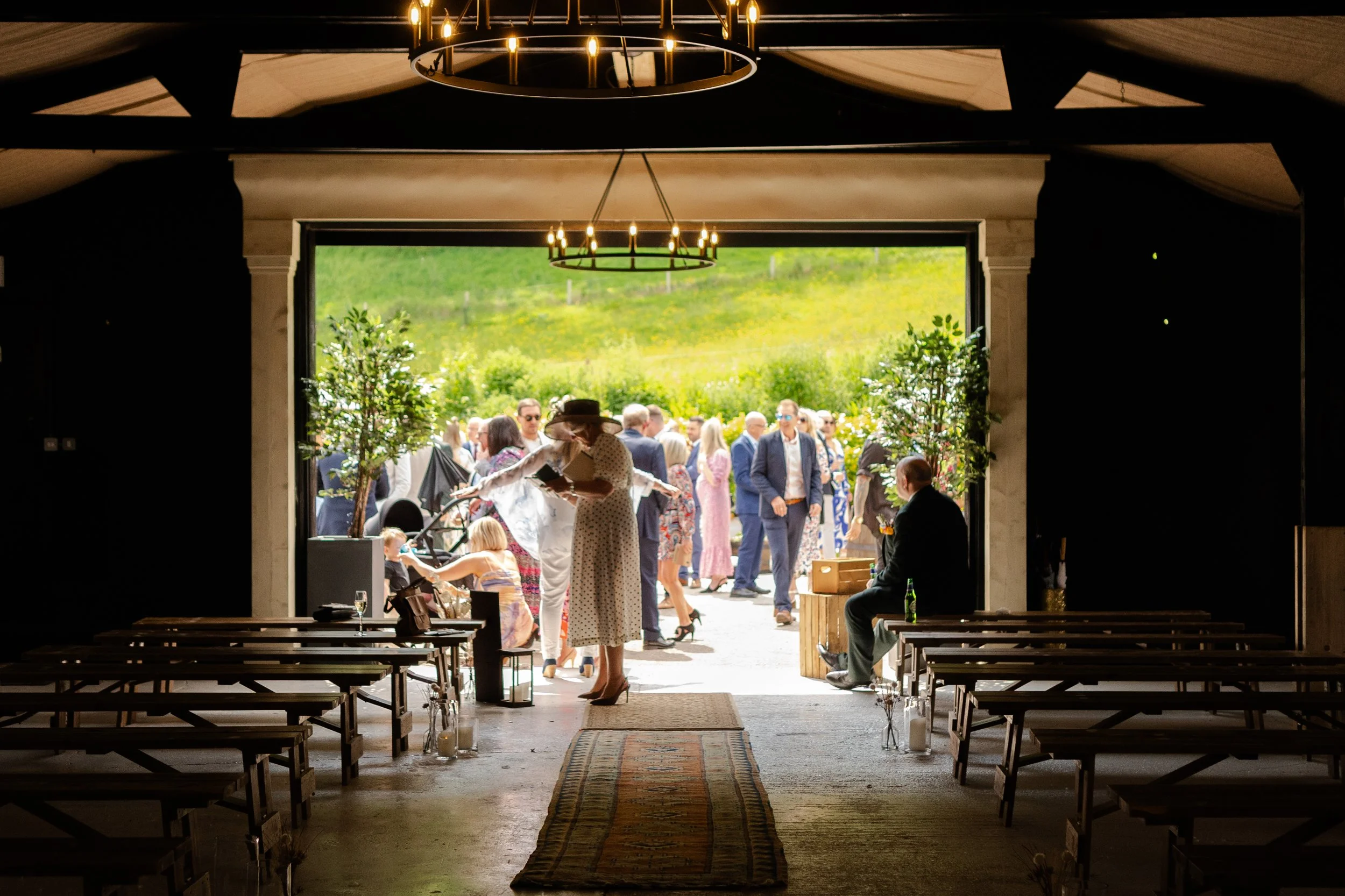 Ceremony room setup in a barn, shot from the front looking down the aisle to outside with guest gathering at a rhyse farm wedding