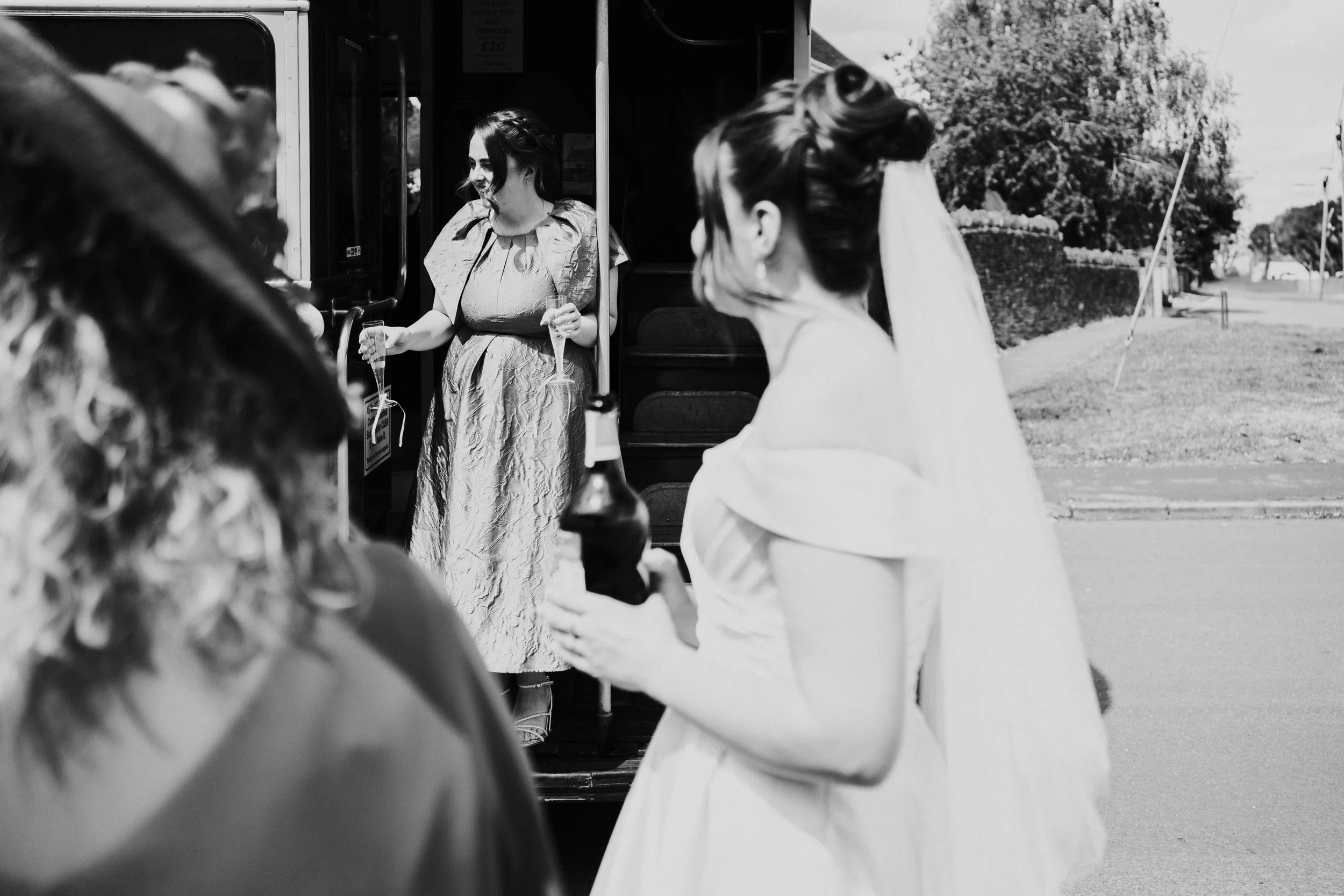 Bride looking at Bridesmaid standing on the routemaster step holding a champagne glass at a Worton Kitchen Garden, Oxfordshire Wedding.