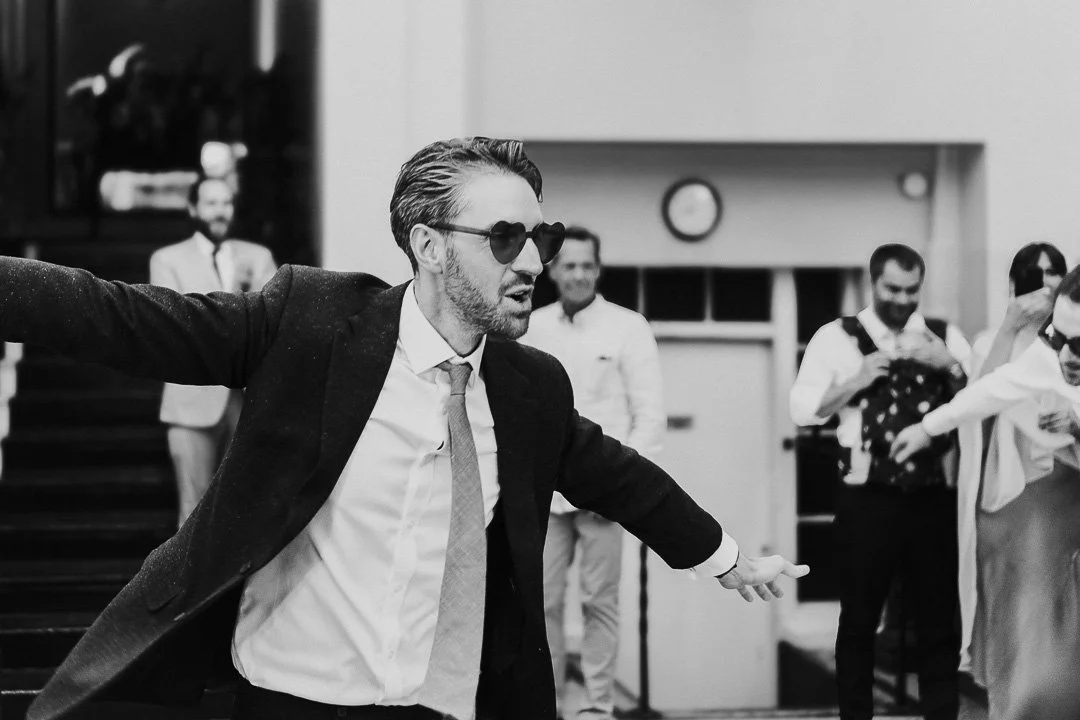 Groom on the dancefloor at Hampton Court House pretending to be flying with arms stretched out wearing heart-shaped sunglasses at a London Wedding.