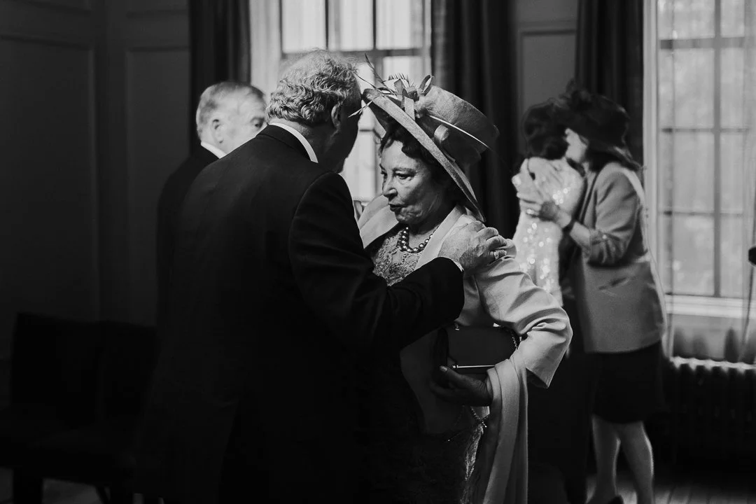 Groom's father knocking Bride's mum large hat off after kissing her on the cheek in the ceremony room at the Old Marylebone Town Hall Wedding.