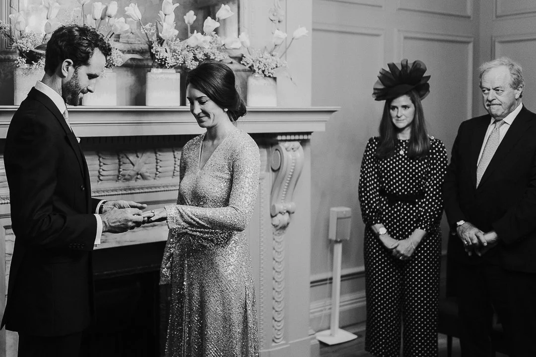 Couple holding hands, both looking at Bride's ring while Groom's Father and Sister look on, in the ceremony room at the Old Marylebone Town Hall.