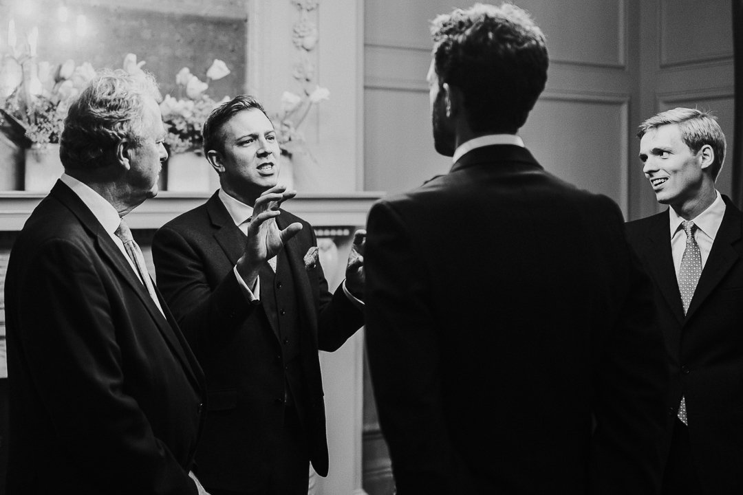 Guests speaking to Groom & other guests while making hand gestures in the ceremony room at the Old Marylebone Town Hall Wedding.