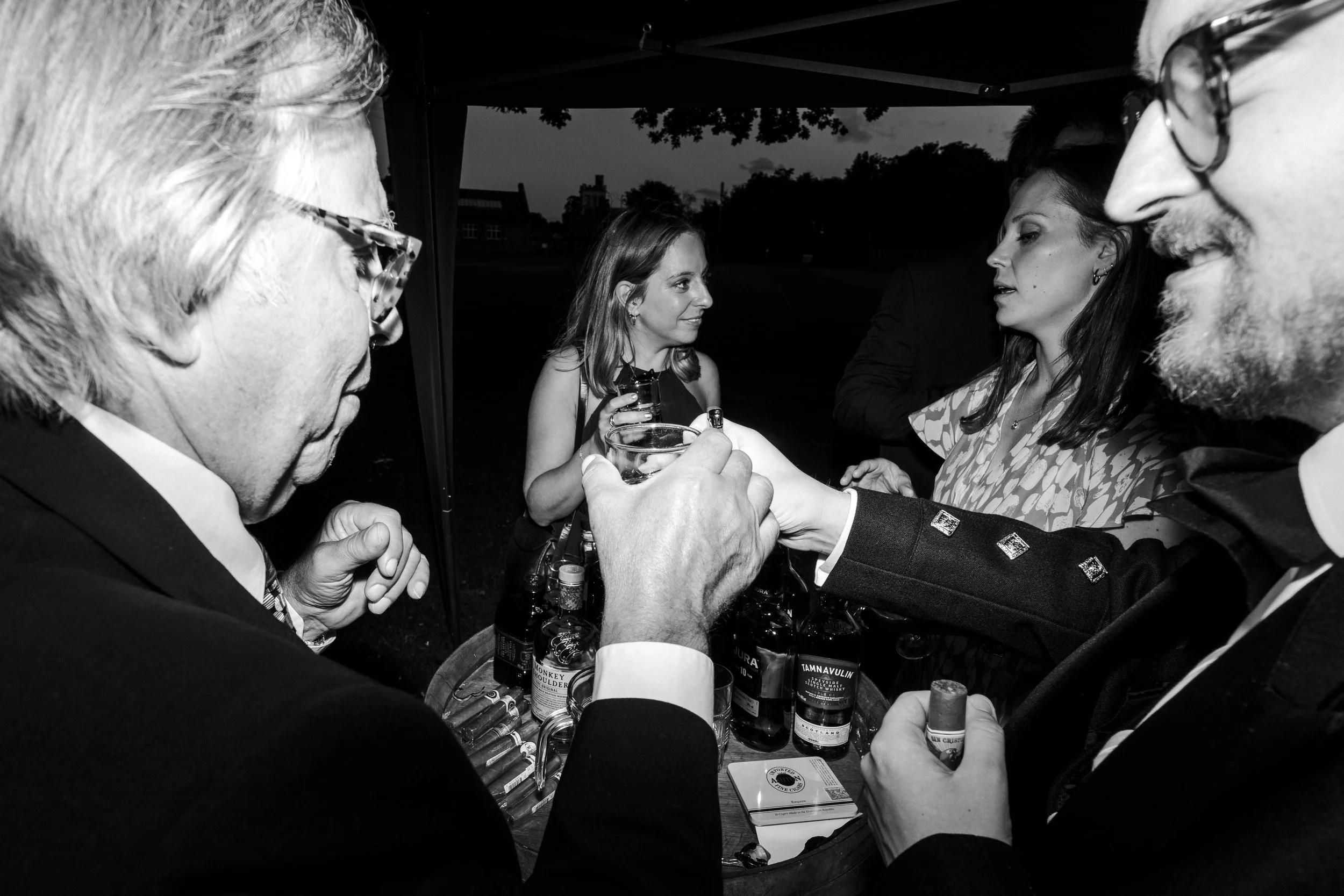 Two wedding guests toasting each other under canopy set up as Whiskey and Cigar Bar at a Highgate, London Wedding.