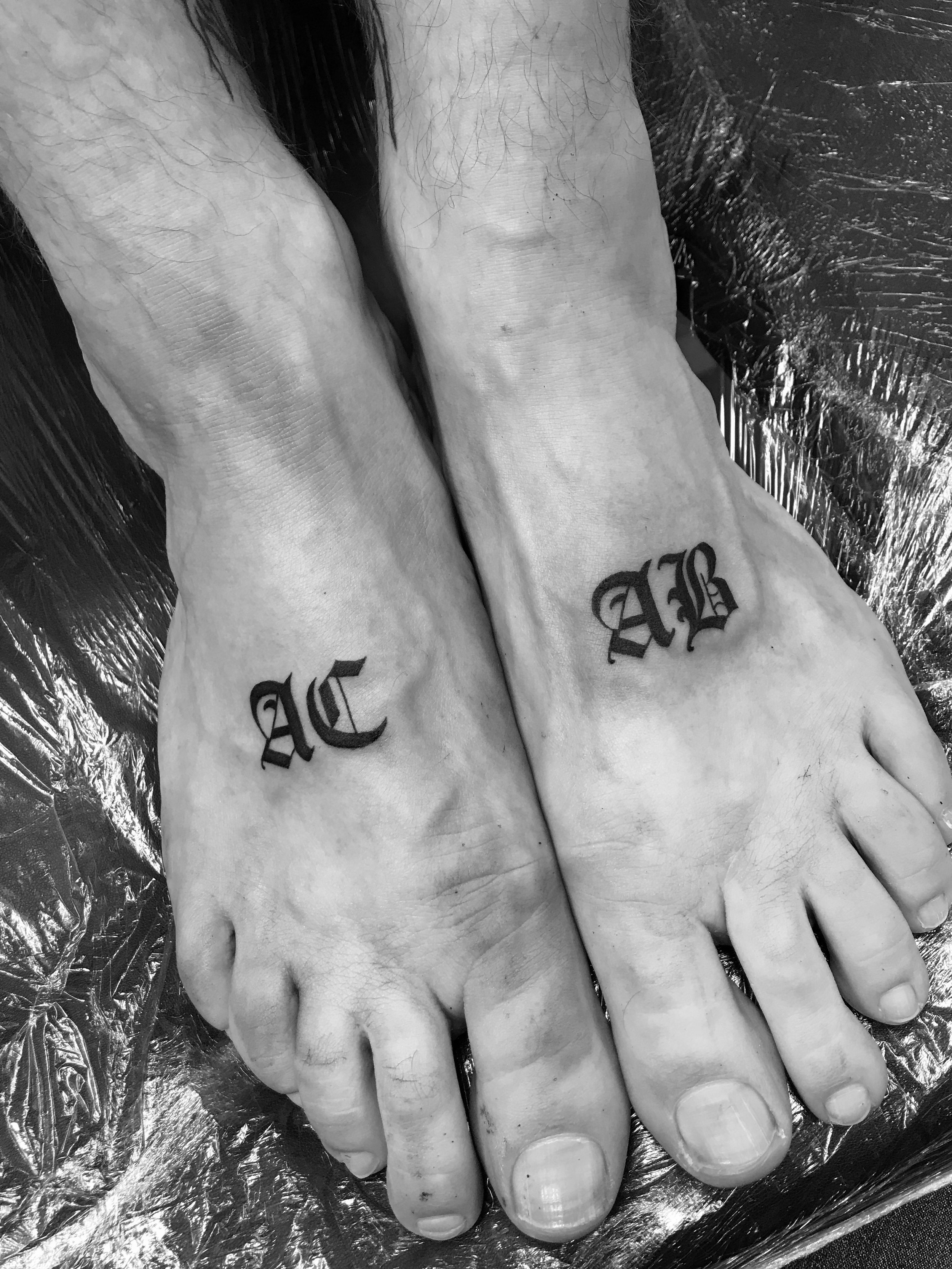 Black and white photo of two feet with tattoos "AC" on the left foot and "AB" on the right foot.