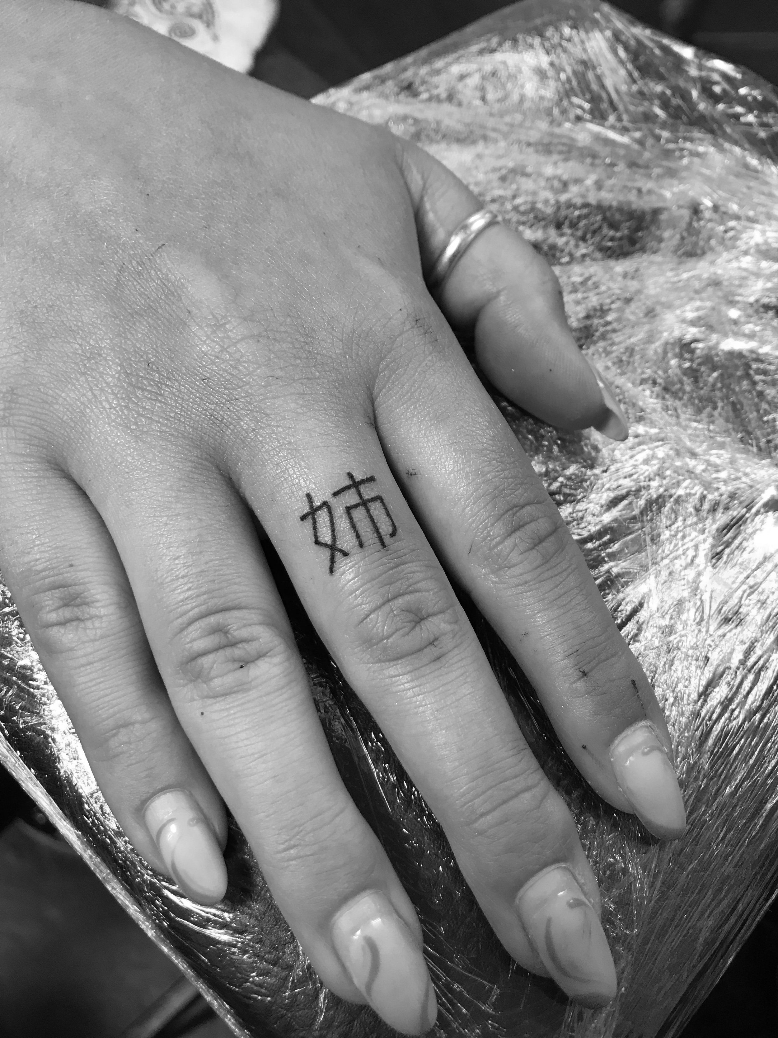 Black and white image of a hand with manicured nails and a ring on the thumb. The finger has a tattoo of Chinese characters.