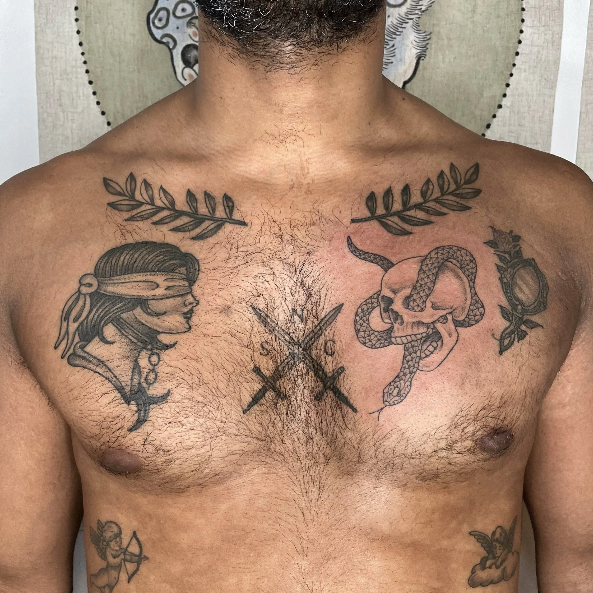 Close-up of a man's chest tattoos, featuring a blindfolded woman, a skull with a snake, crossed swords with letters, olive branches, a rose, and a cherub with a bow and arrow.