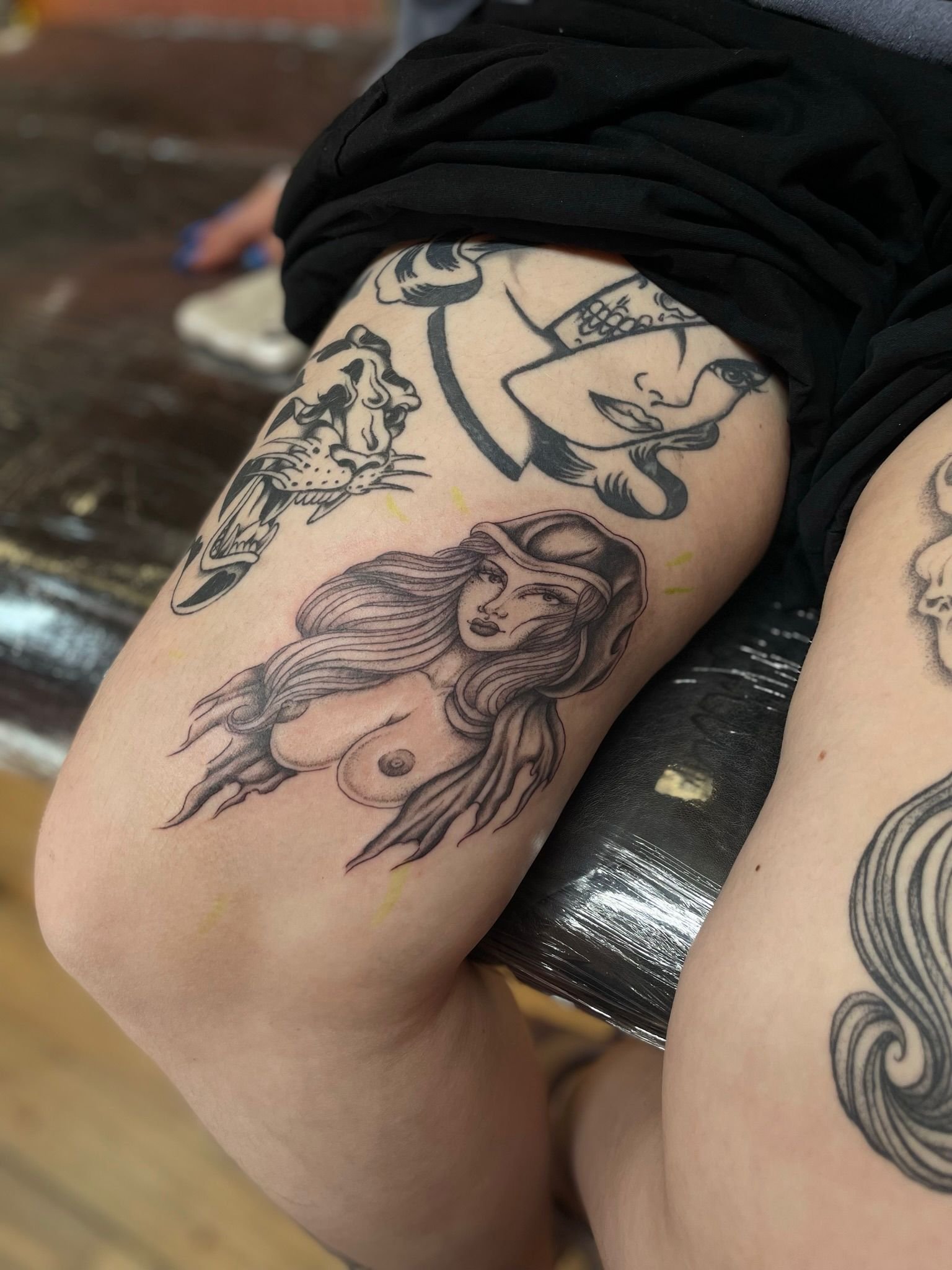 Tattoo art on thighs featuring a woman's portrait, a roaring panther, and a woman with skeletal features.
