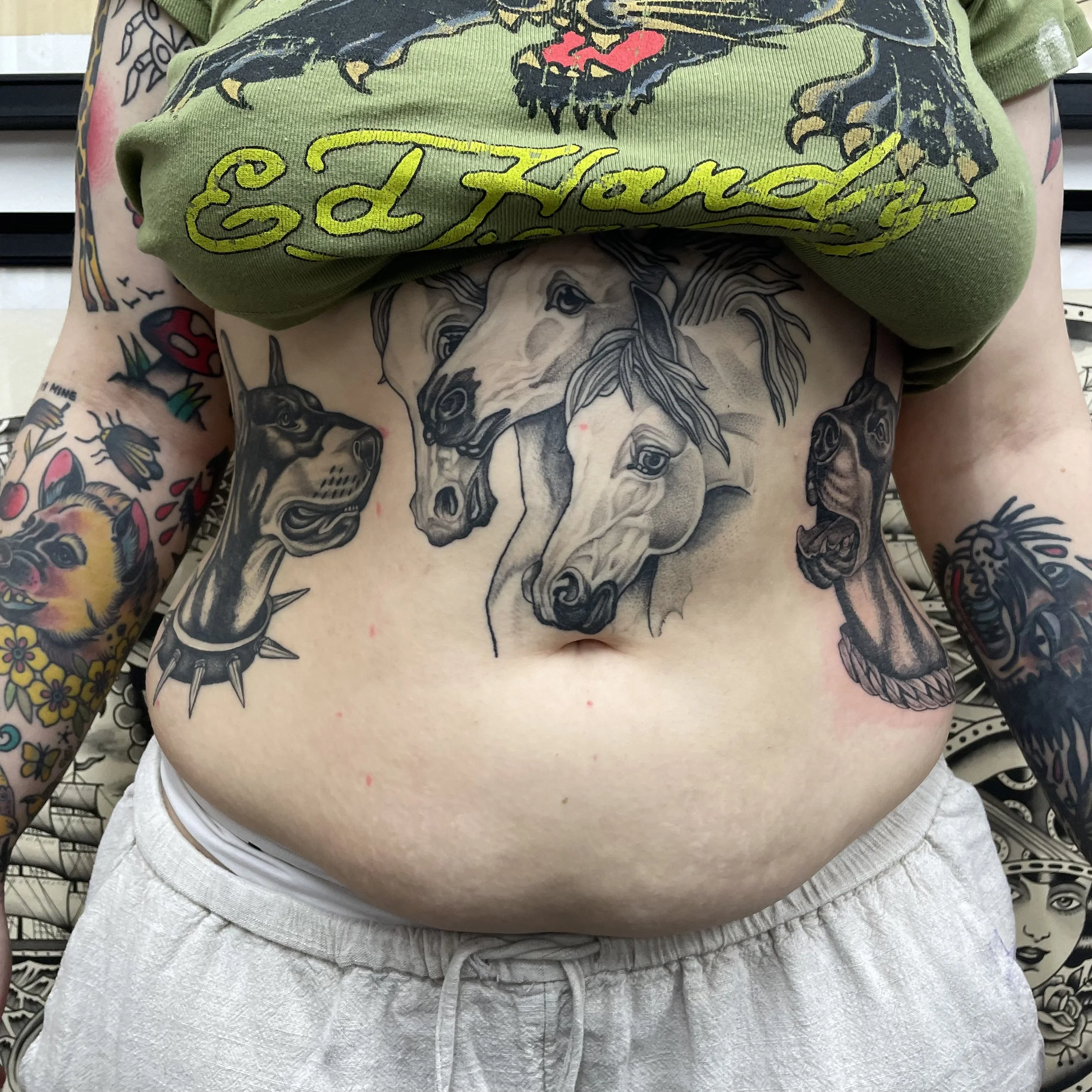 Person showing tattoo of horses on their stomach with additional tattoos of dogs and flowers on their arms, wearing an Ed Hardy shirt and light-colored sweatpants.