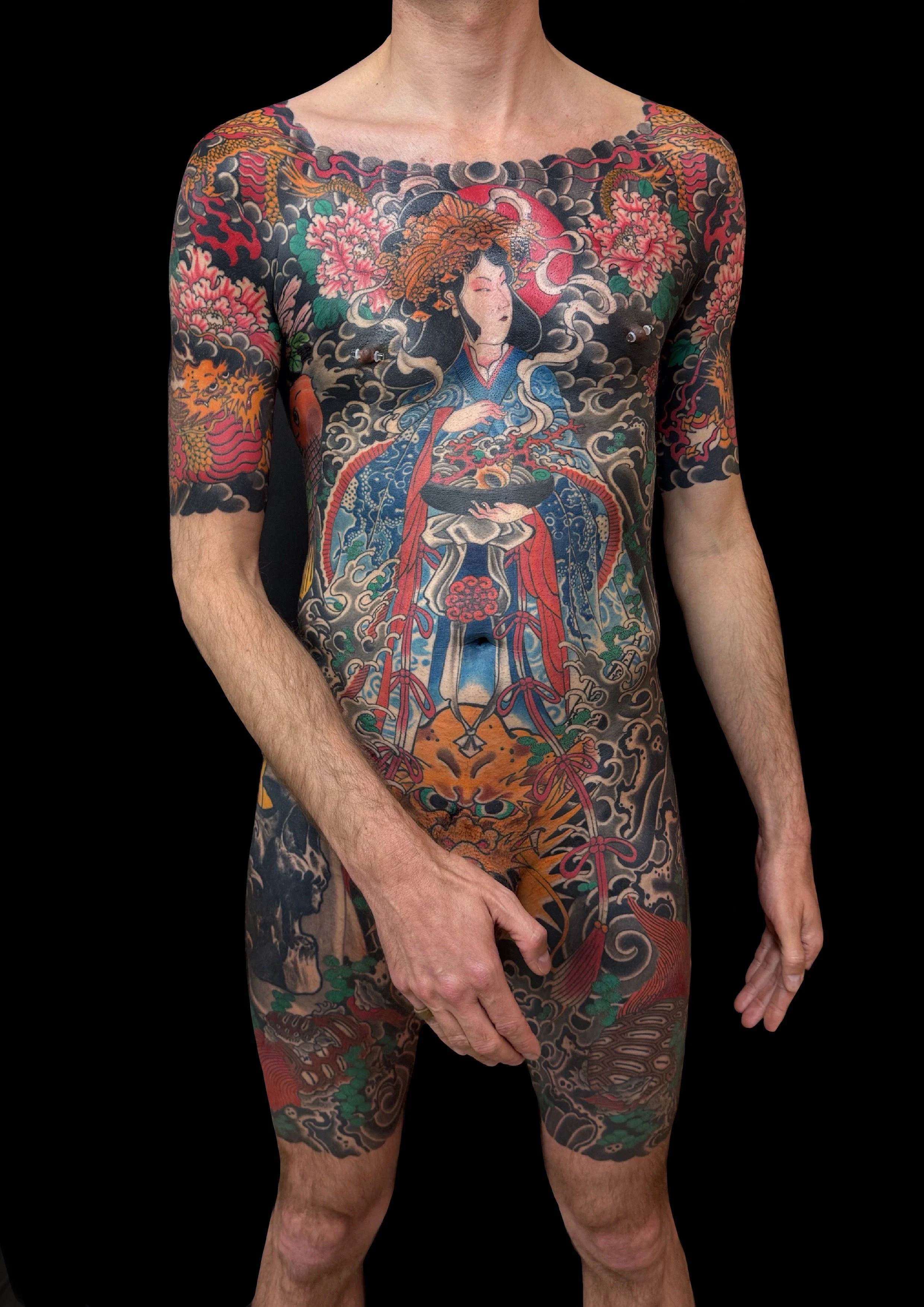 A person with a full-body traditional Japanese style tattoo, featuring intricate designs of a figure in elaborate dress, surrounded by floral motifs, clouds, and mythical elements, covering the torso, arms, and legs.