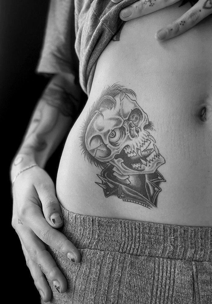 Person showing a black and gray tattoo of a skull with hair, wearing a jacket, on their side torso near the waistline.