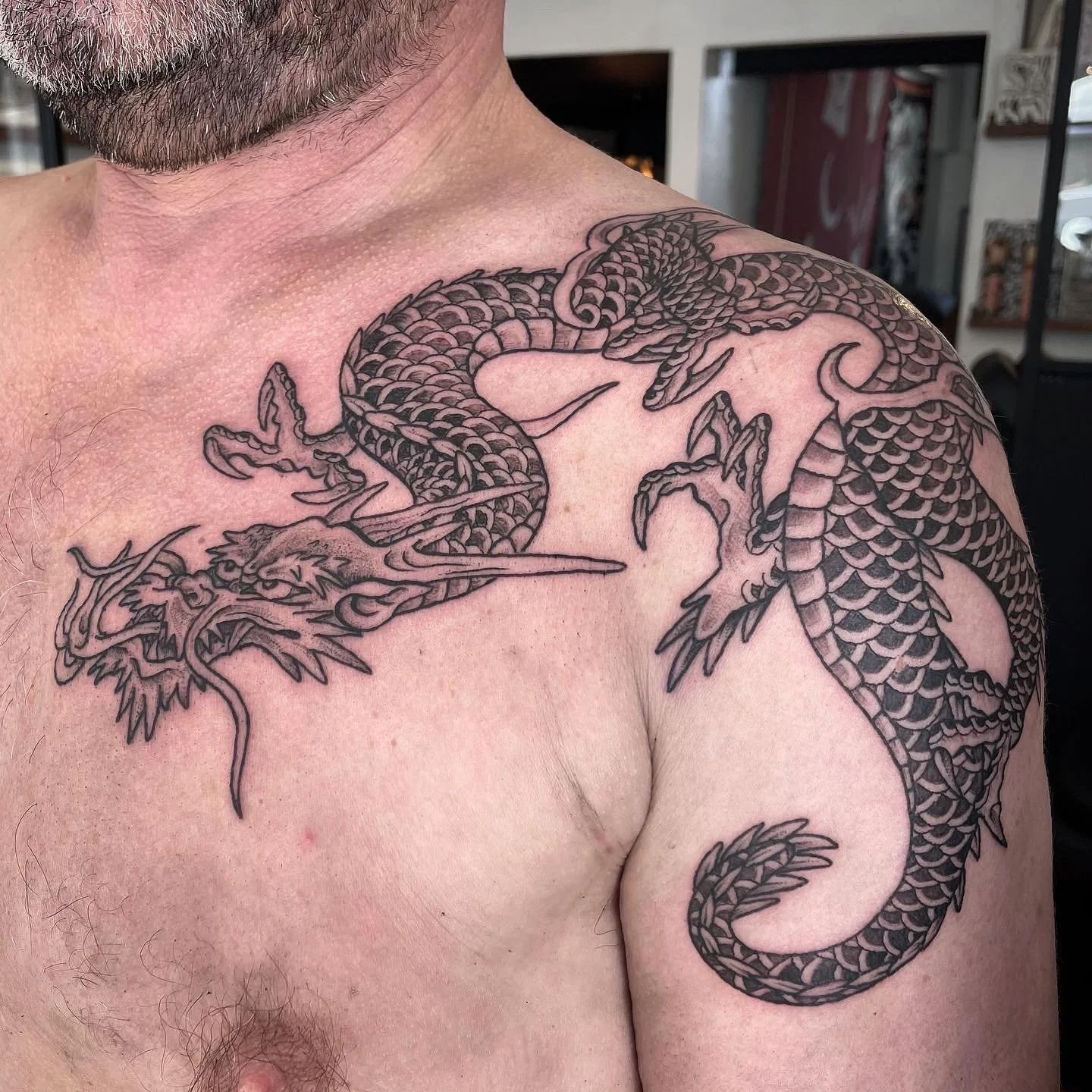 Close-up of a man’s shoulder and chest with a black and gray dragon tattoo, featuring detailed scales, claws, and facial features, wrapping around the shoulder.