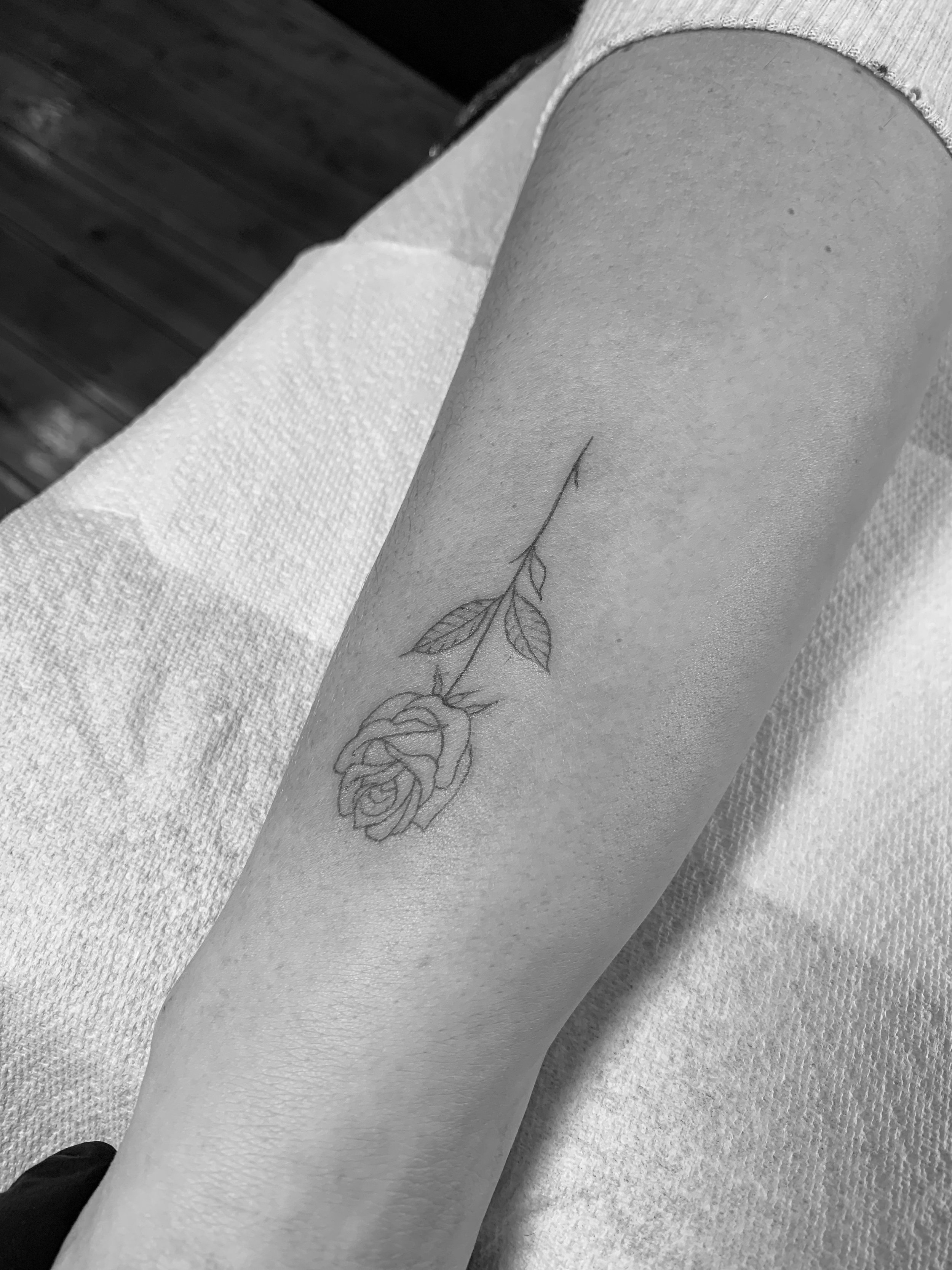 Black and white tattoo of a rose with stem and leaves on a forearm.