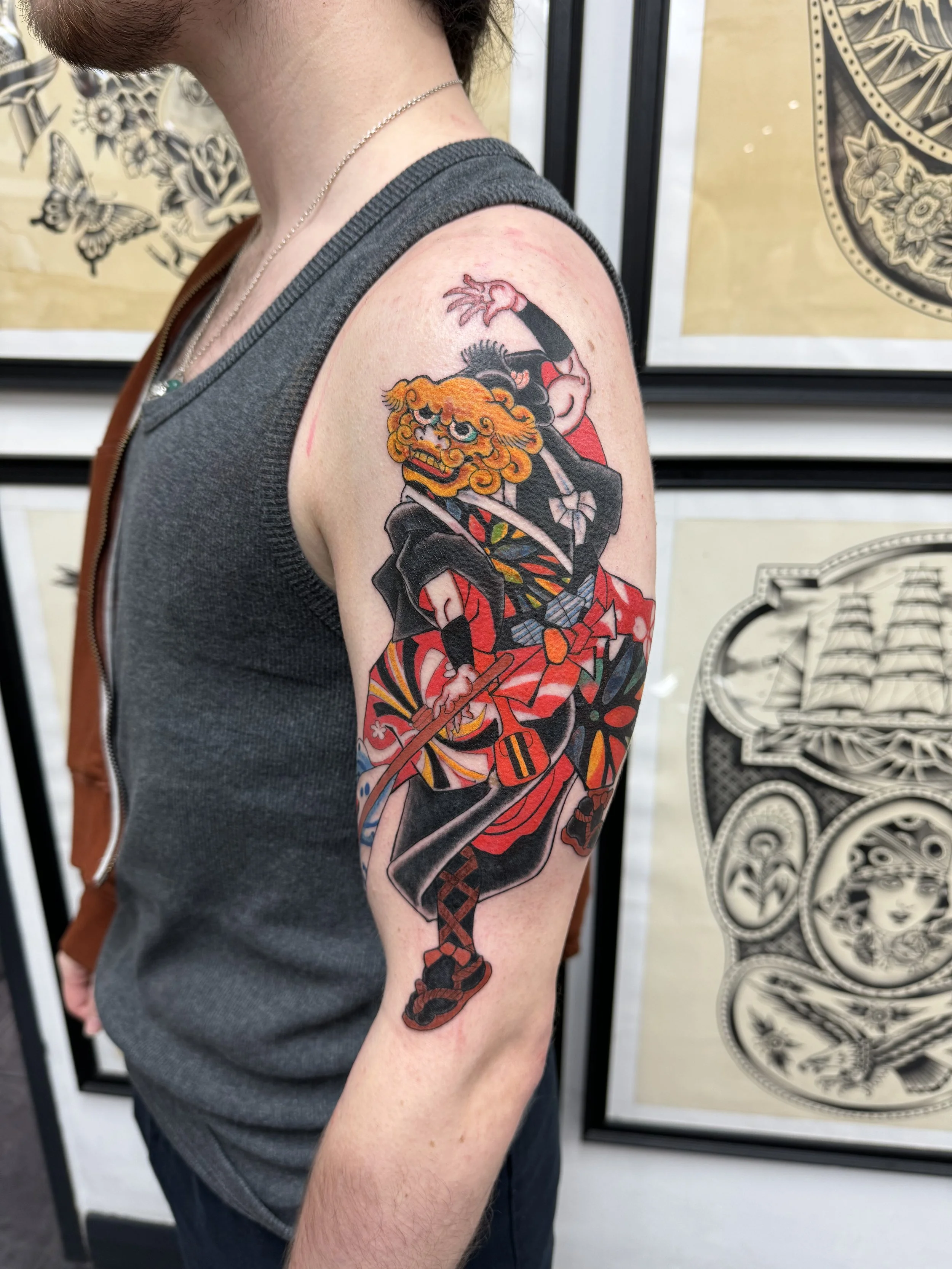 Person with a colorful tattoo depicting a Japanese demon or oni wearing a traditional outfit, on their upper arm. Background shows framed artwork on the wall.