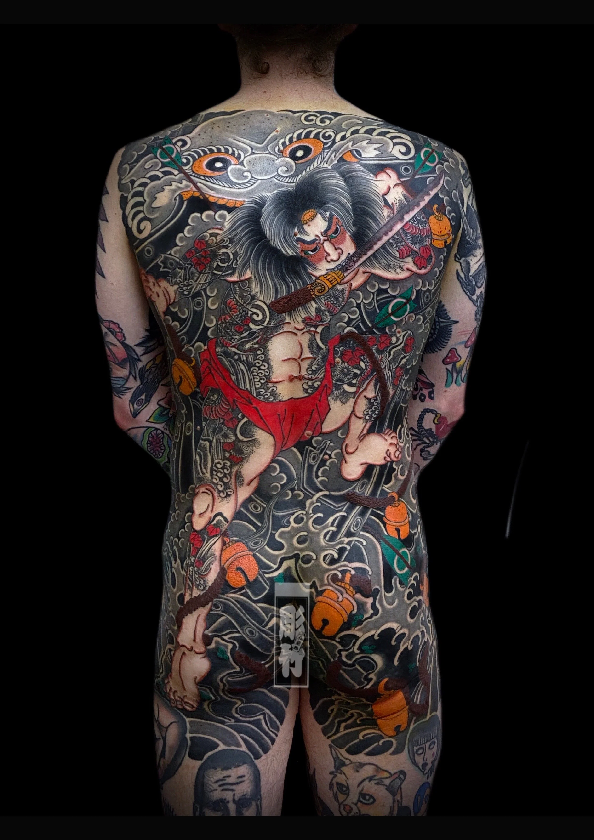 Back view of a person with an elaborate full-body tattoo featuring a traditional Japanese theme, including a warrior, waves, and various mythical elements, on a black background.