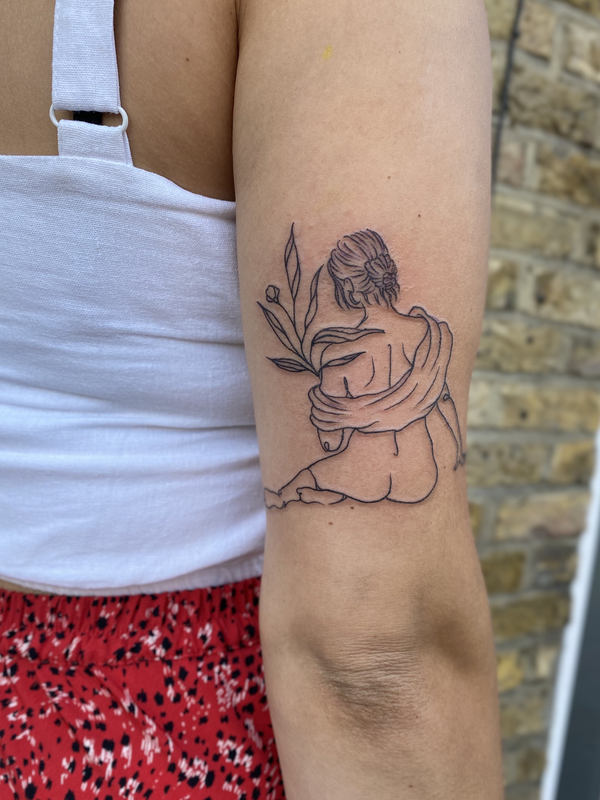 Tattoo of a line drawing on a person's arm depicting a woman sitting with her back turned, holding a flower with leaves.