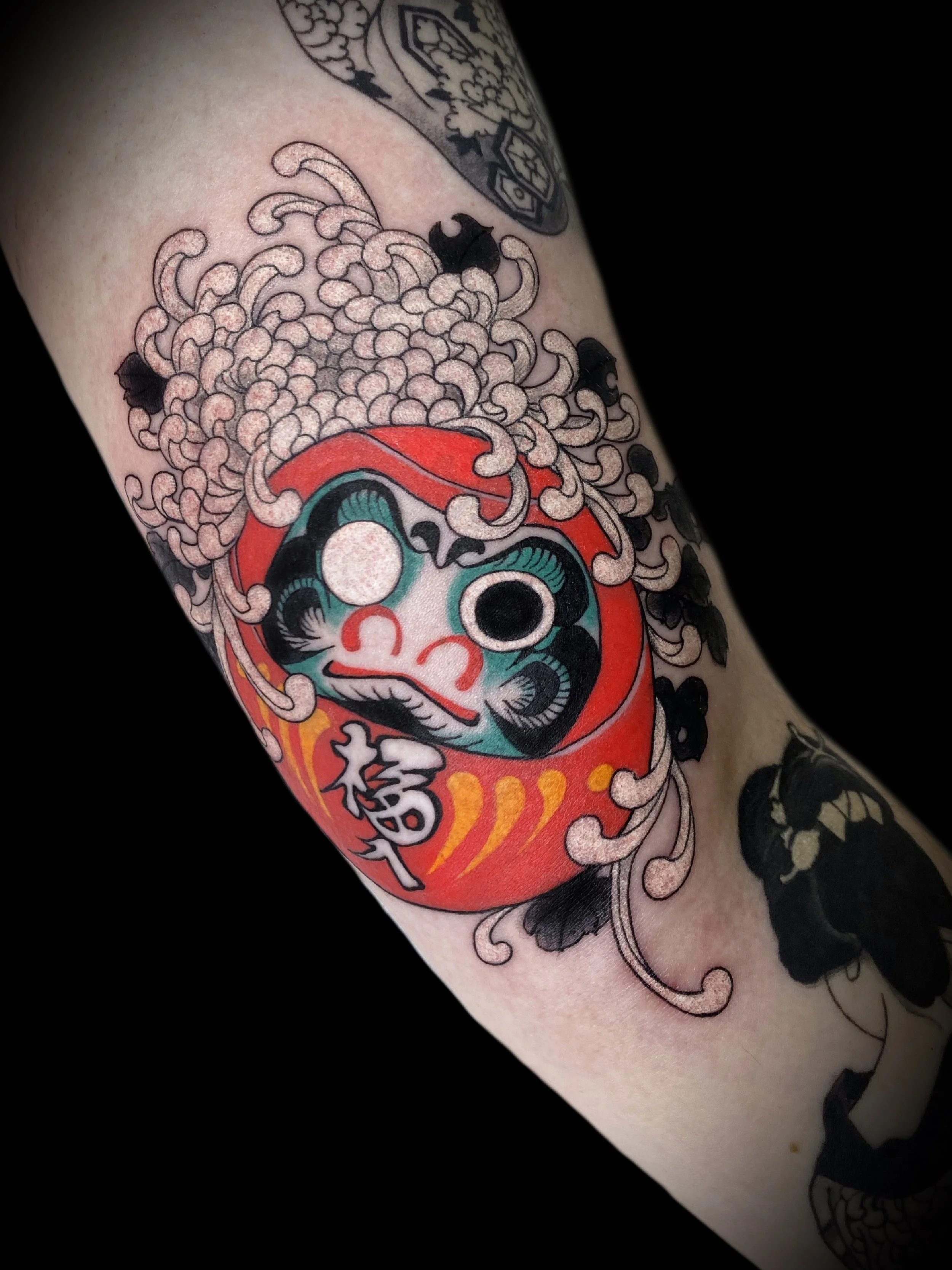Tattoo of a red Daruma doll with stylized, swirling designs and Japanese characters on an arm.
