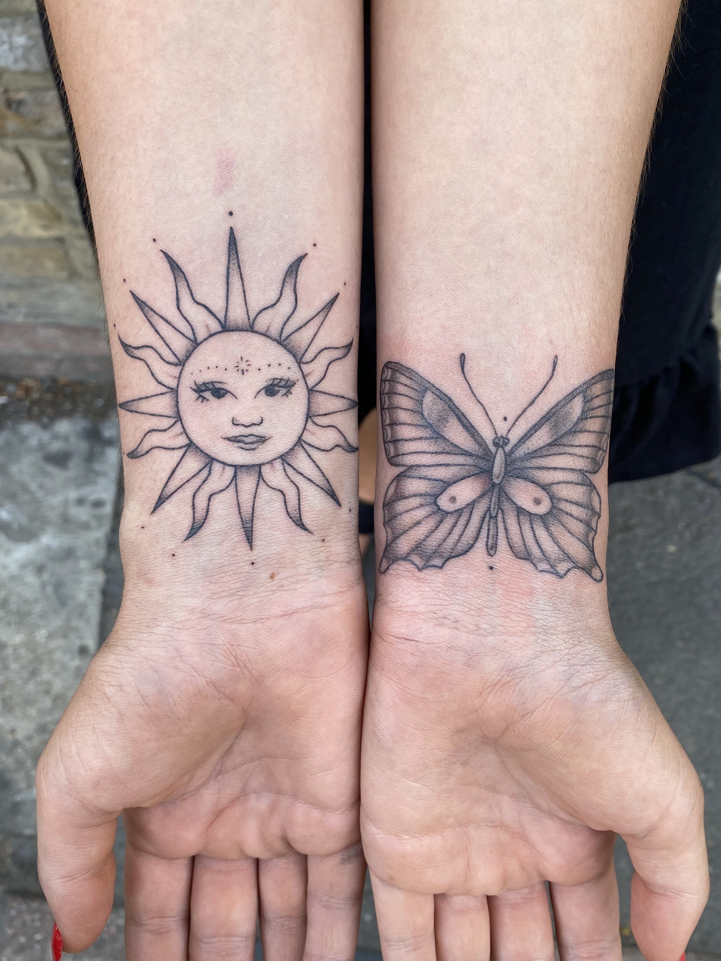 Tattoo of a sun with a face on one wrist and a butterfly on the other wrist on a person's forearms.