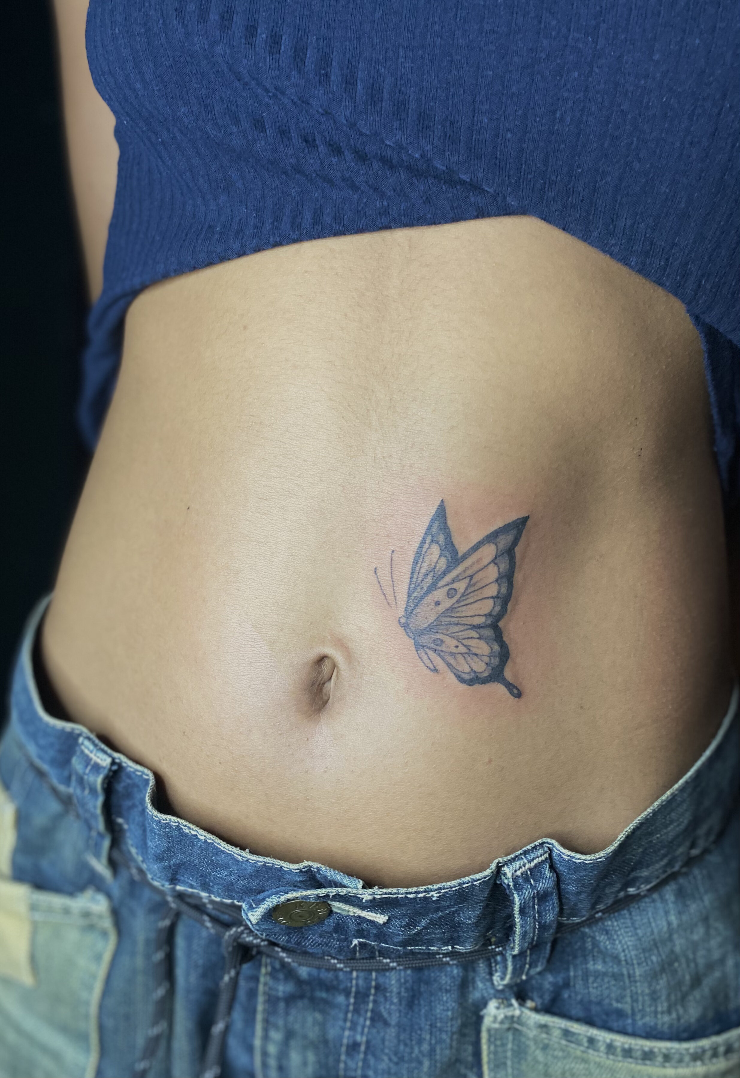 Close-up of a person's lower abdomen with a butterfly tattoo near the belly button, wearing blue jeans and a blue top.