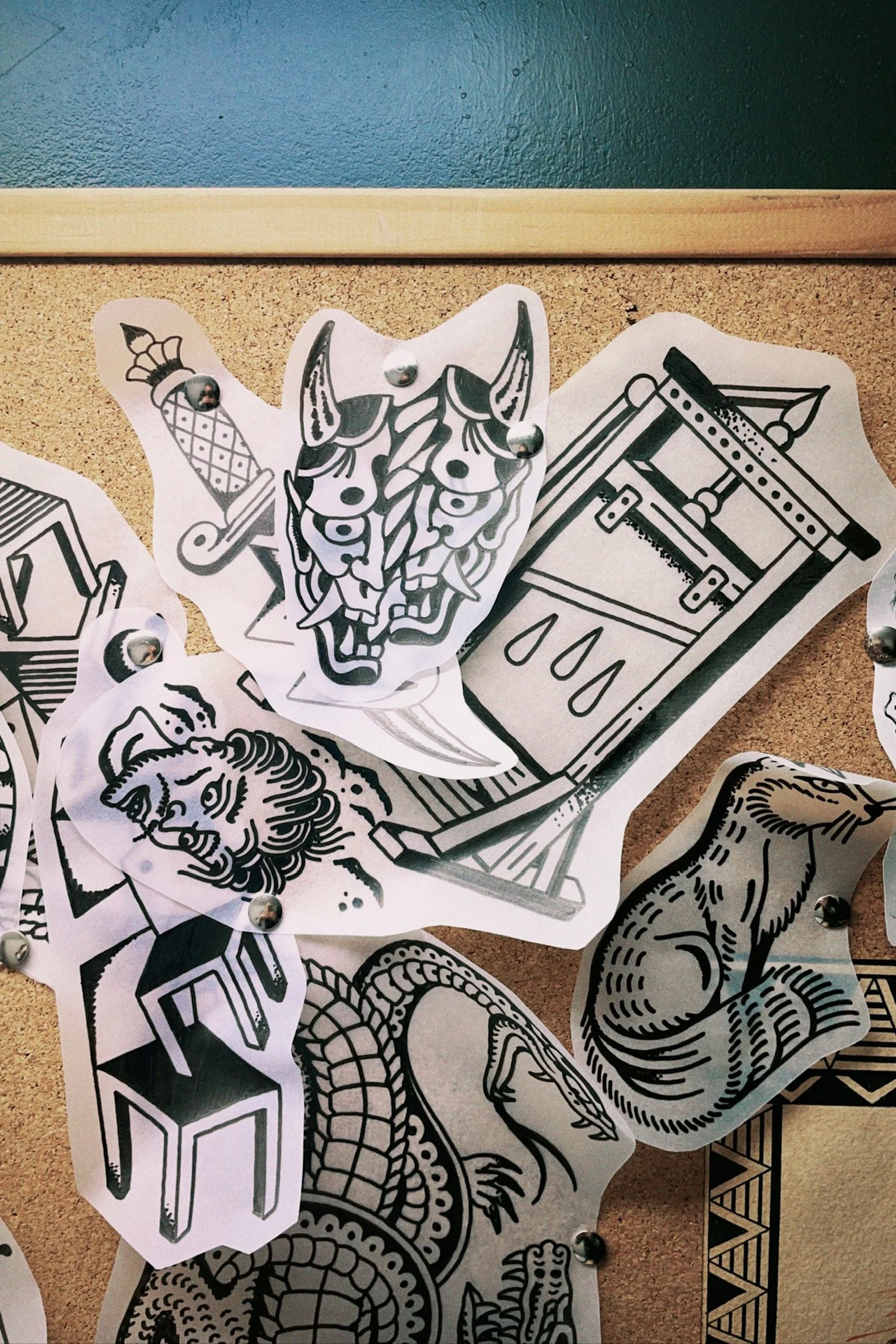 Tattoo flash sheet on corkboard featuring black and white designs including a demon mask, guillotine, chair, crocodile, and seal.