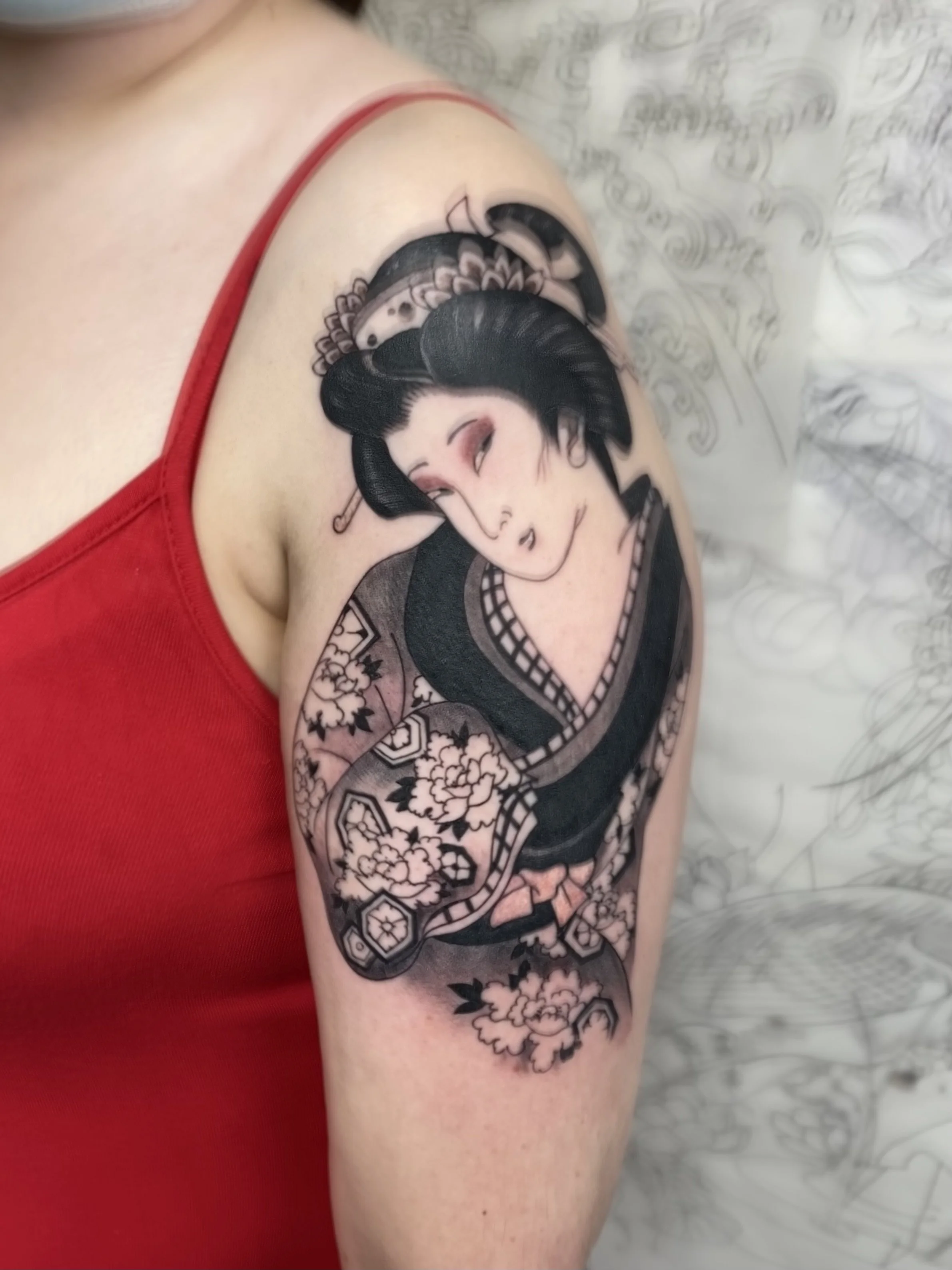 Tattoo of a geisha on a person's upper arm, wearing a red tank top.