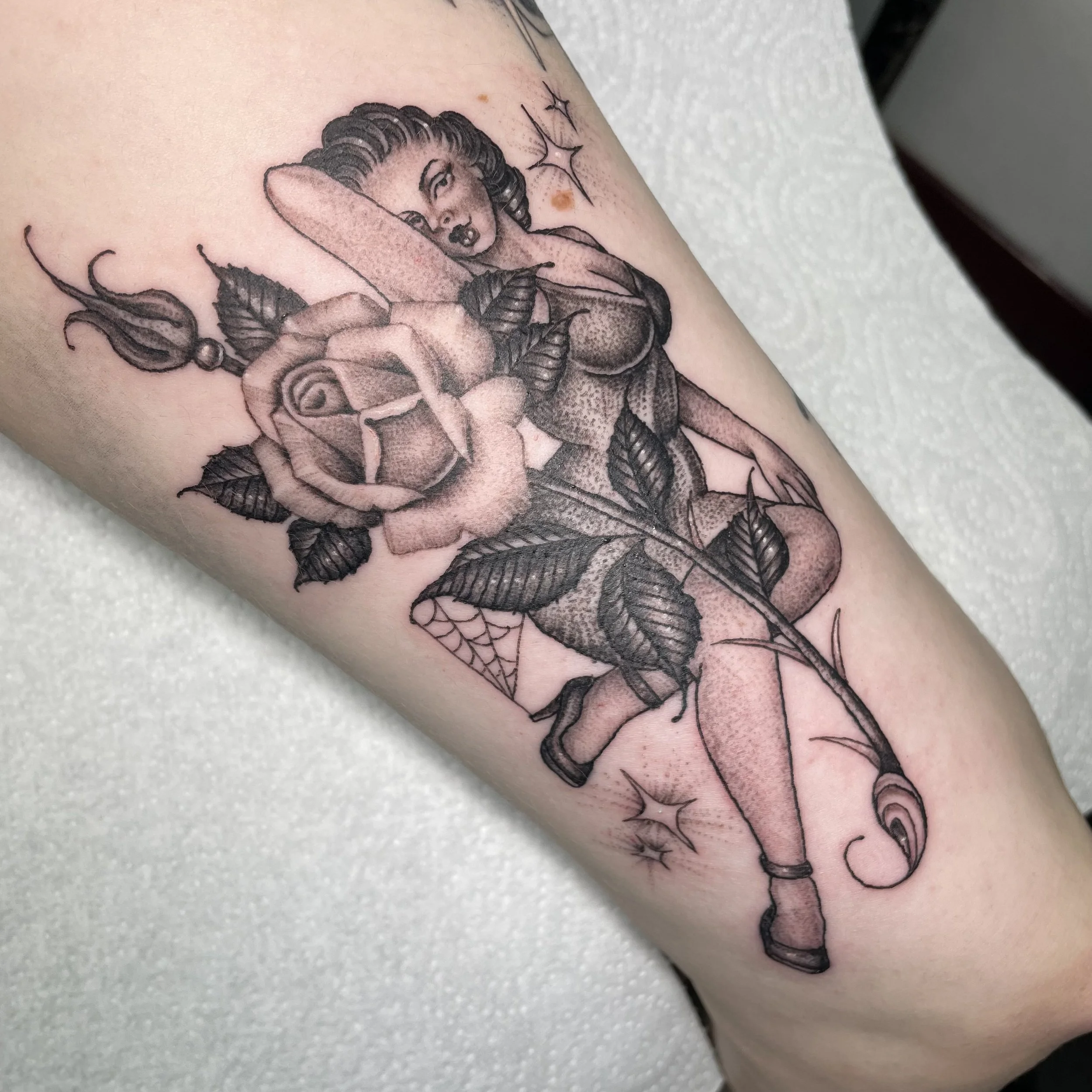 Tattoo of a pin-up style woman with a large blooming rose, leaves, and thorned stem integrated with the design. The woman appears to be stepping forward, surrounded by decorative elements such as stars and web-like patterns.