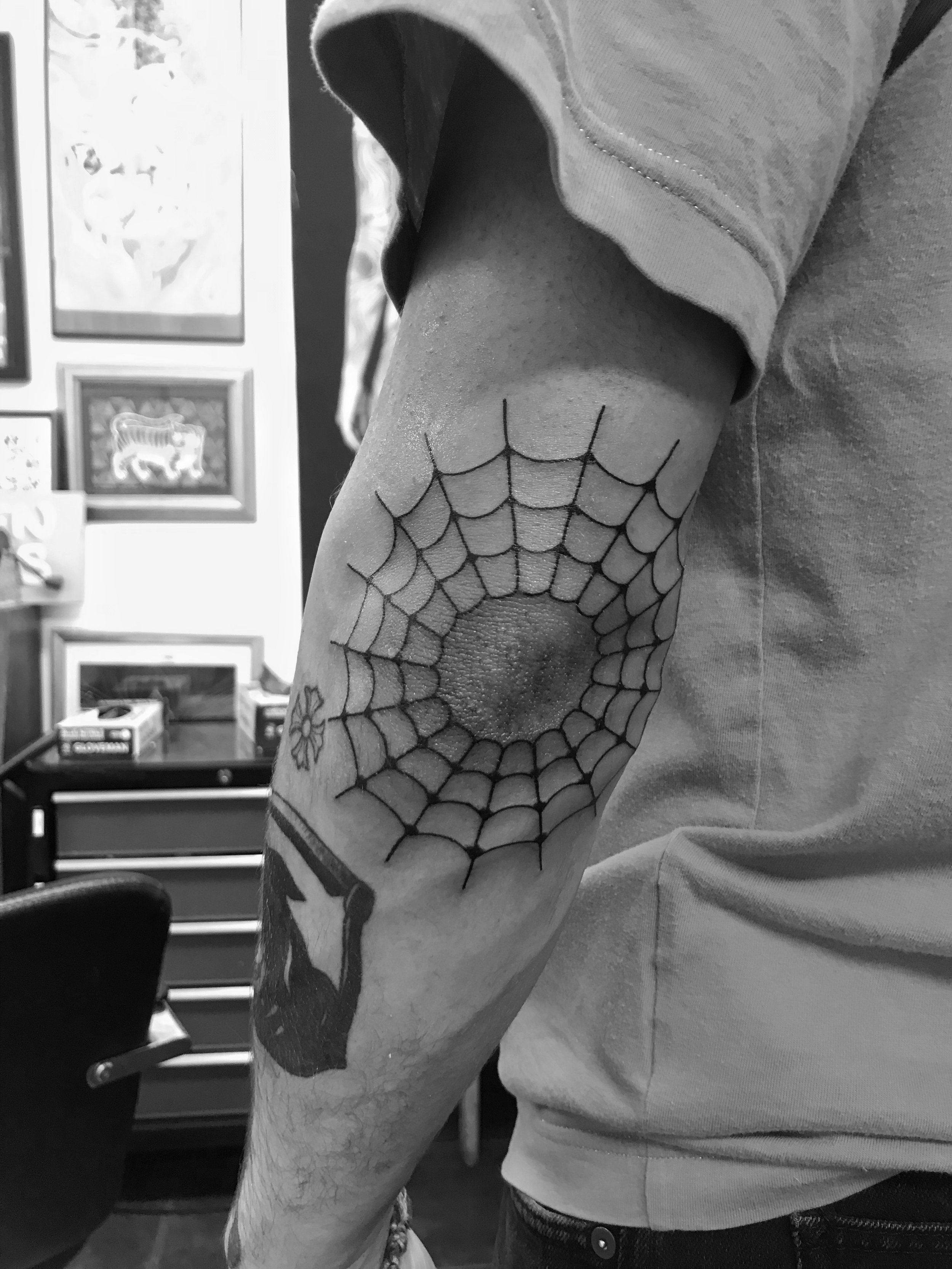 Tattoo of a spider web on a person's elbow, with additional arm tattoos. Black and white photo.