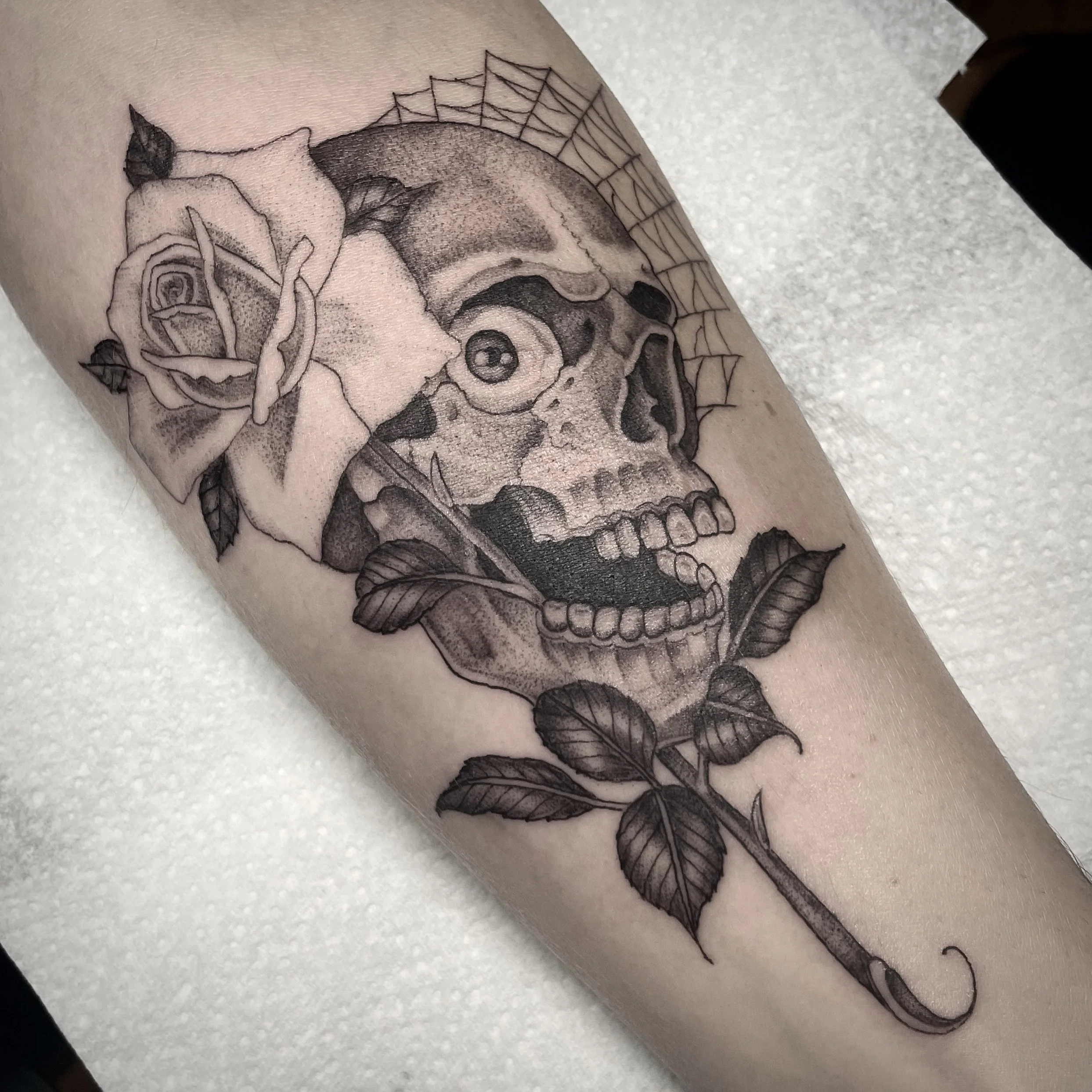 Tattoo of a skull with a rose on a person's arm. The skull is detailed with a cobweb, and the rose is depicted in full bloom with leaves.