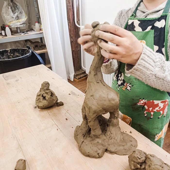 Person modelliert eine große Tonfigur an einem Arbeitstisch in einer Keramikwerkstatt.