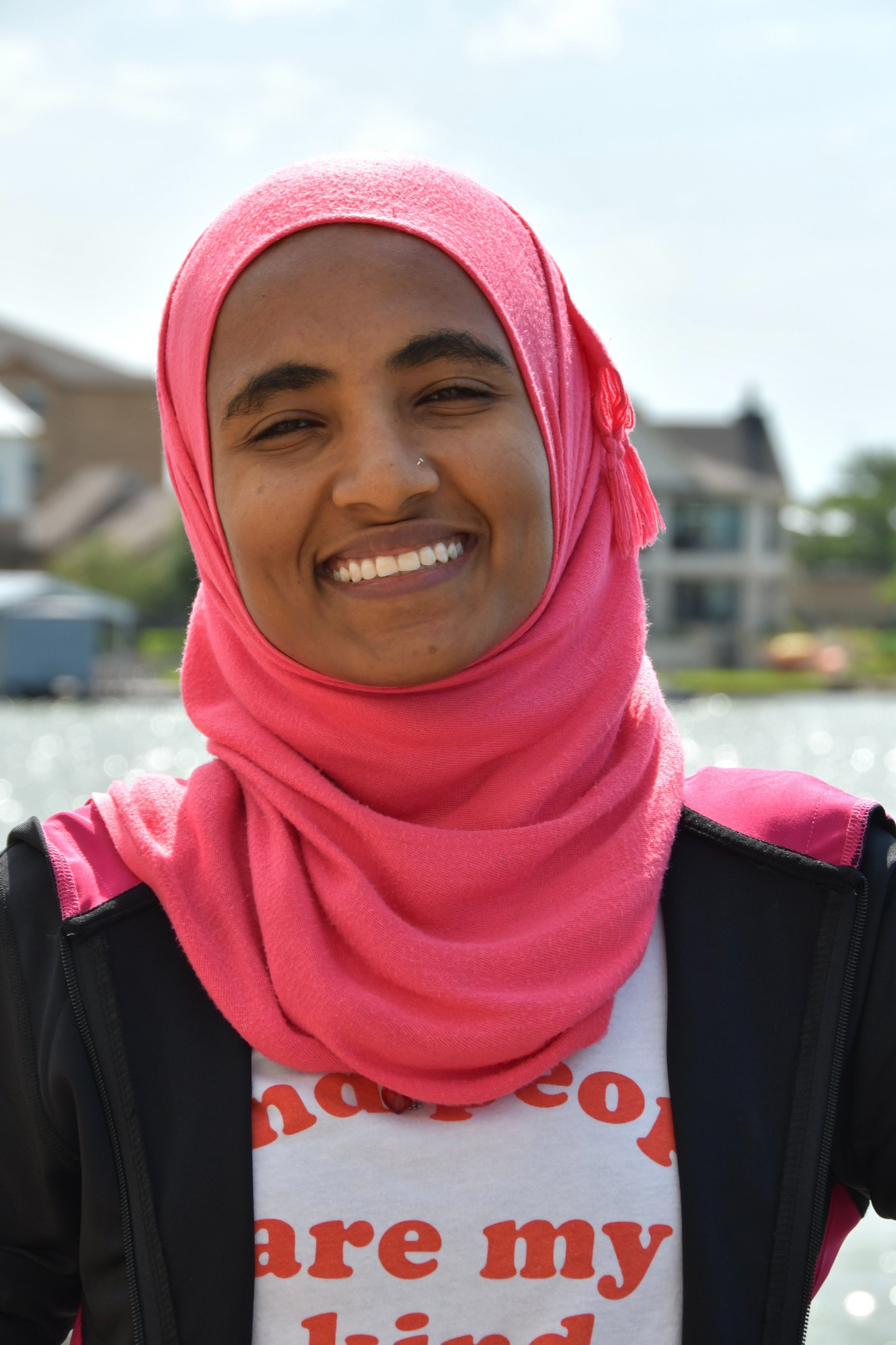 Smiling person in a pink hijab near water with houses in the background.