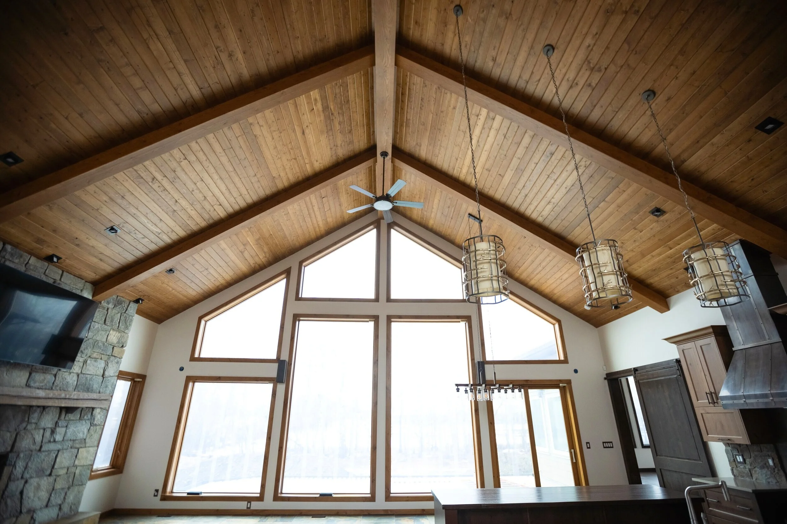 Interior of a spacious room with a high vaulted wooden ceiling, large triangular windows, a ceiling fan, and hanging pendant lights.