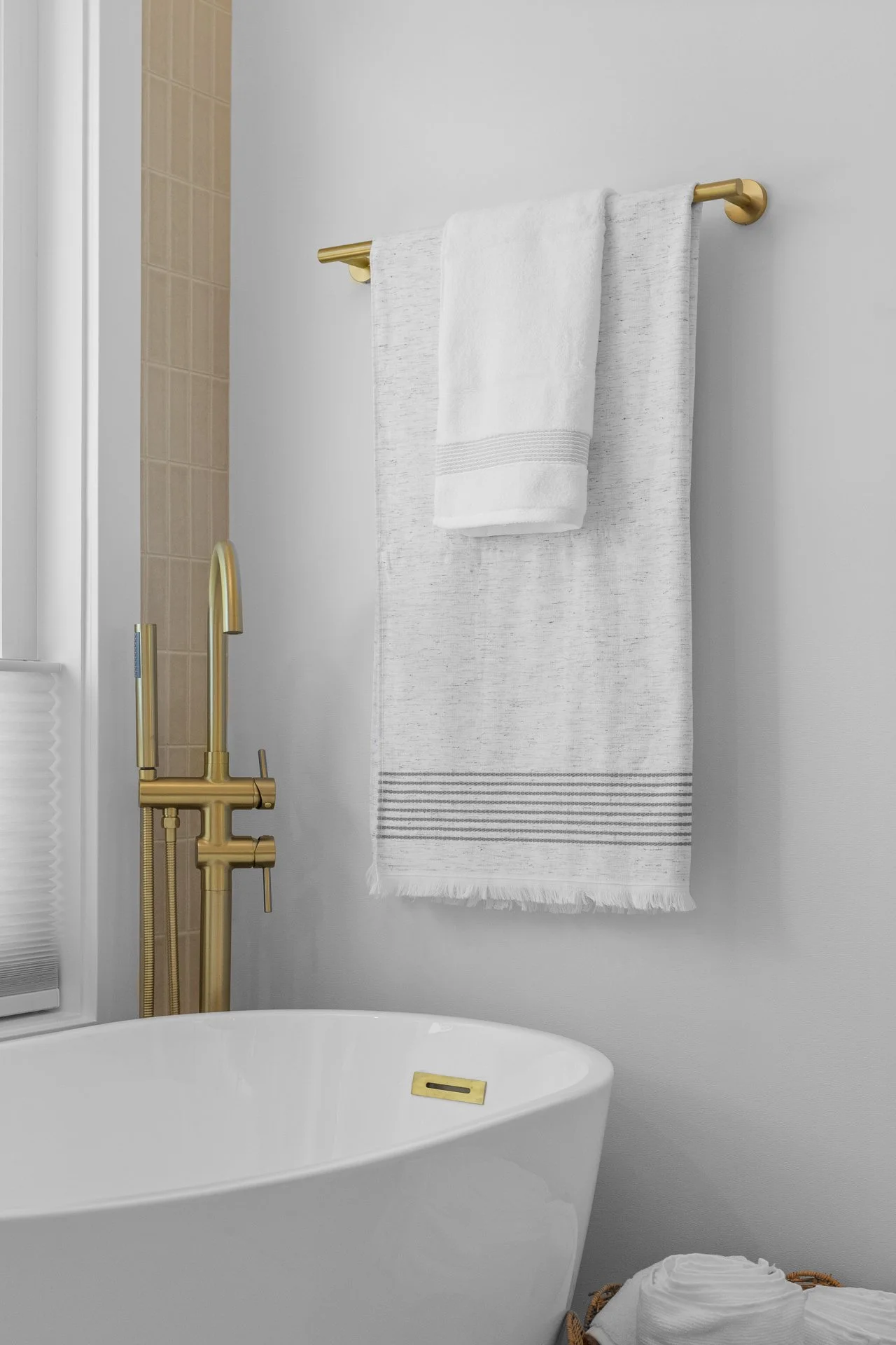 A bathroom with a white freestanding bathtub, a gold faucet, a white towel hanging on a gold towel bar, and a beige shower area visible to the left. A woven basket with rolled white towels is near the tub.