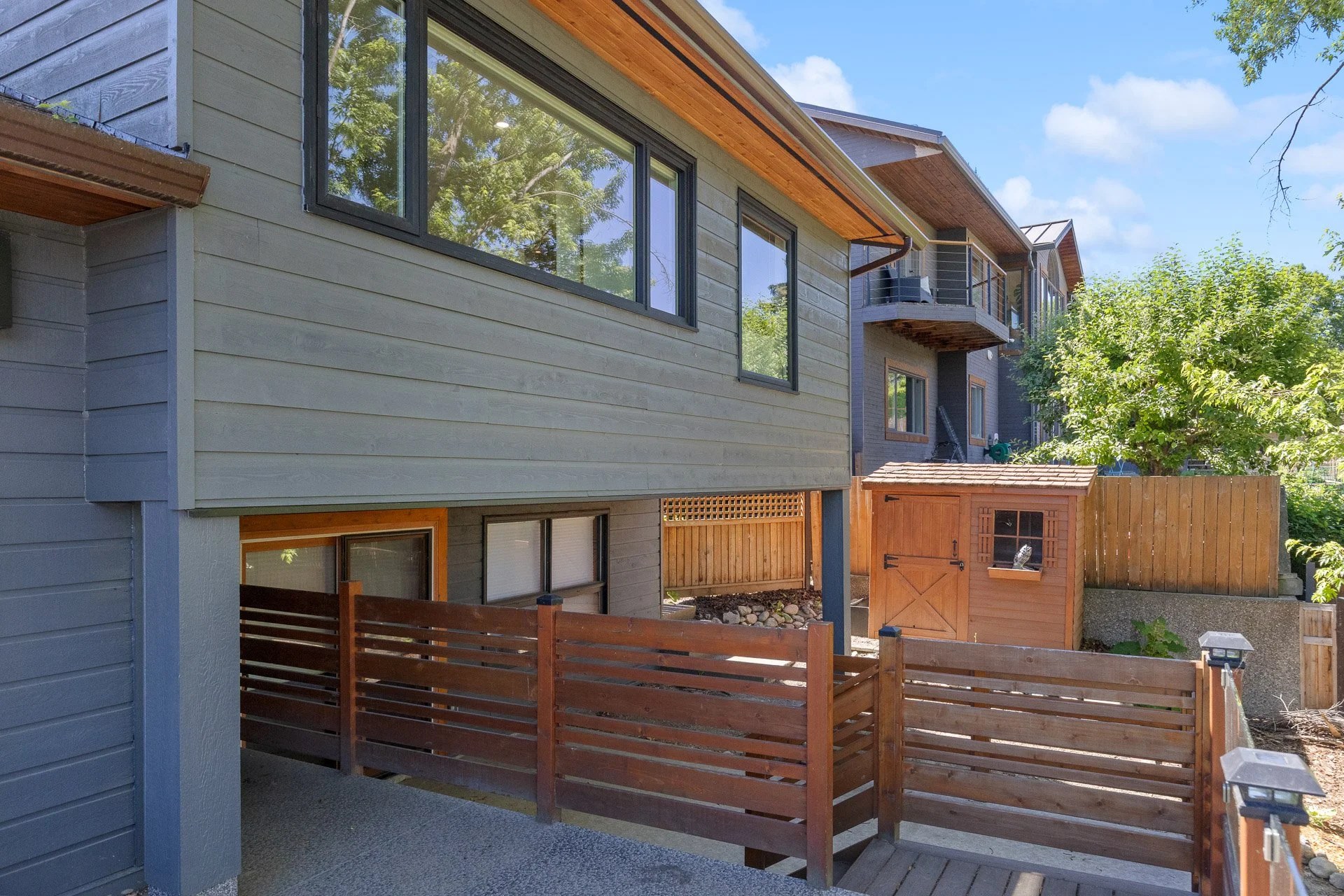 Backyard view of a modern two-story house with gray wooden siding, large windows, and a small wooden shed. A wooden fence surrounds the yard with a tree and blue sky in the background.
