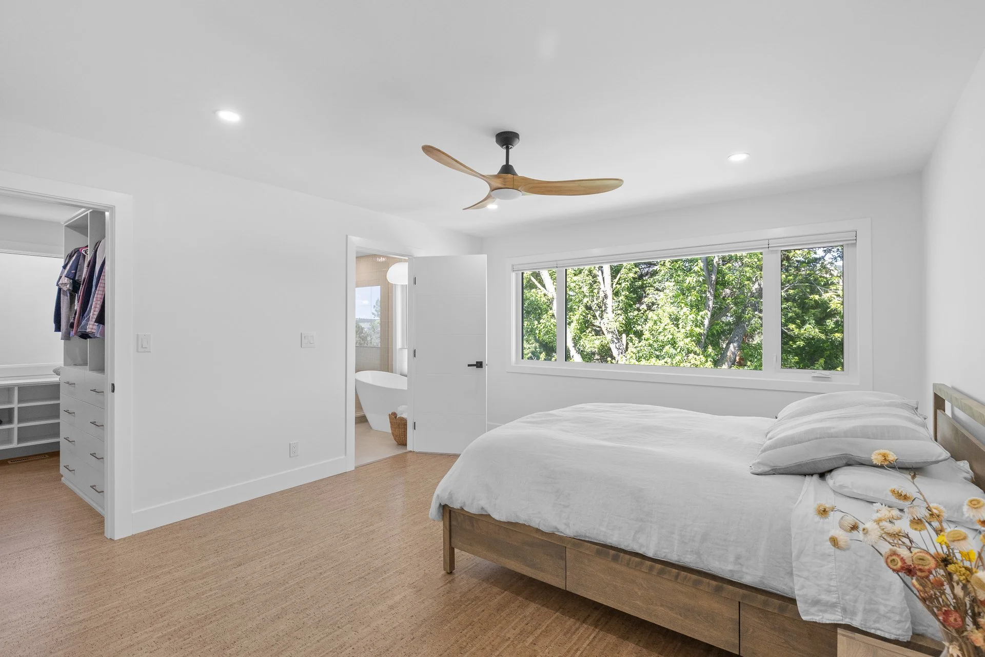 Bright bedroom with white walls, a large window showing green trees outside, a bed with white linens, and a ceiling fan with wooden blades.
