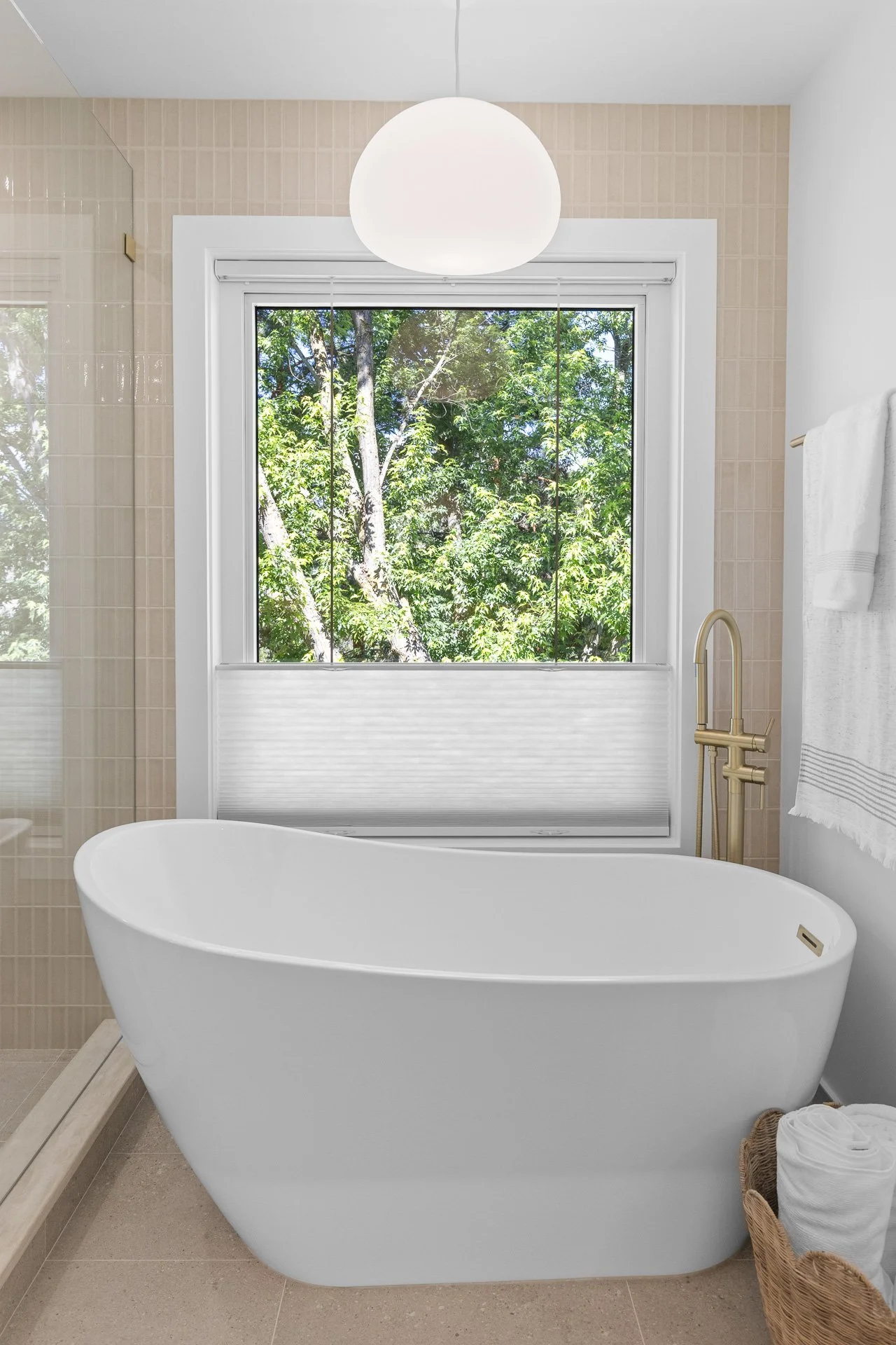 Modern bathroom with a freestanding bathtub, a window showing green trees outside, and beige tiled walls.