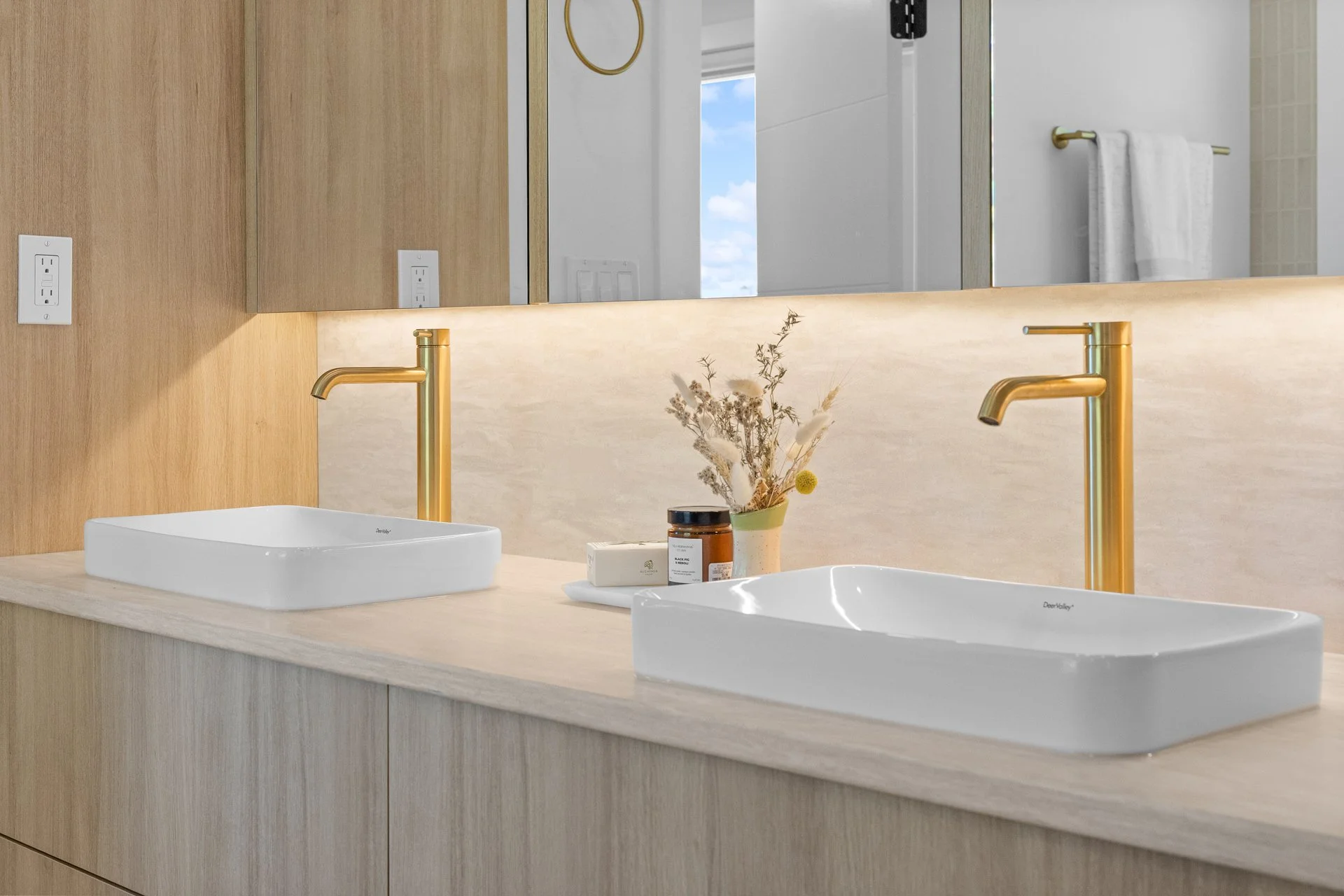 A modern bathroom vanity with dual white sinks, gold faucets, and a large mirror. There are plants and skincare products on the countertop.