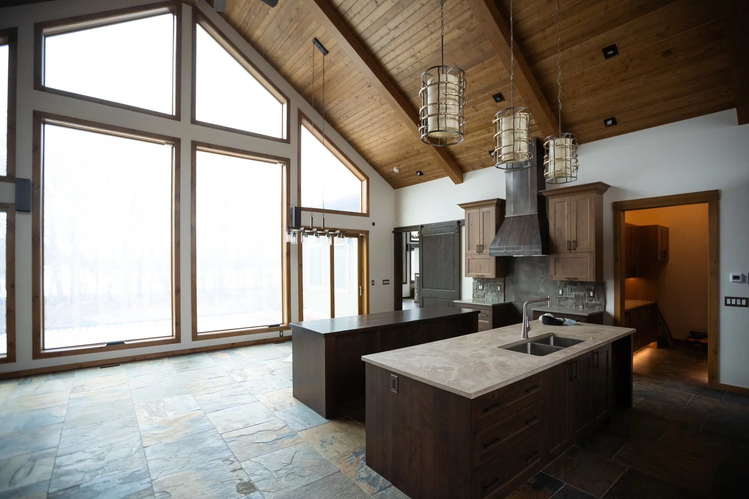 A spacious kitchen with large floor-to-ceiling windows, a wooden vaulted ceiling with pendant lights, a kitchen island with a marble top, and wooden cabinetry.