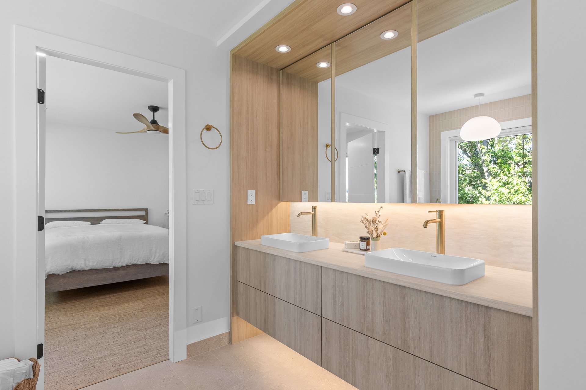 Modern bathroom with double sinks, wooden accents, and large mirror; bedroom with bed and ceiling fan visible through doorway; natural light from window with greenery outside.