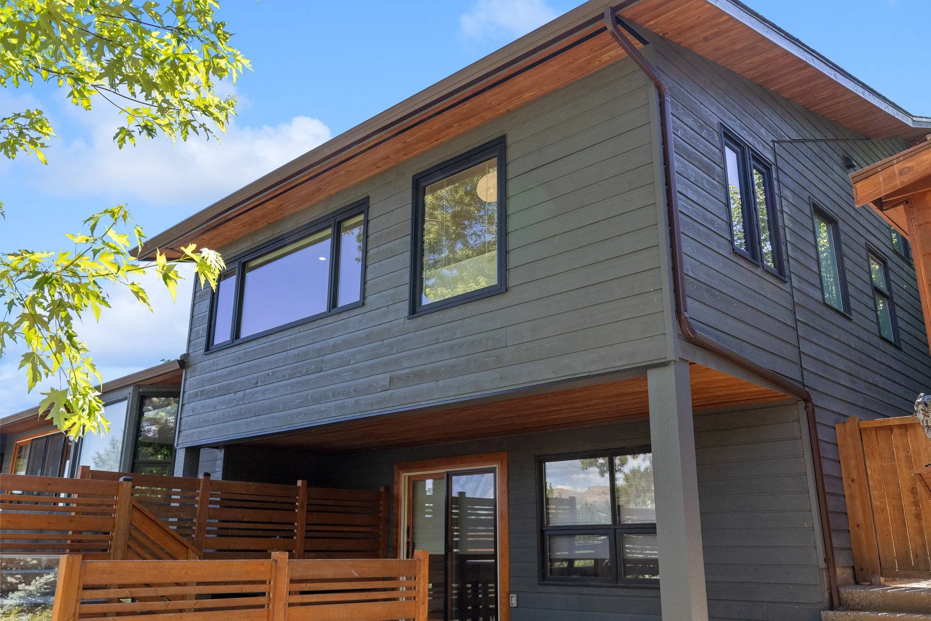 A modern two-story house with gray wooden siding and multiple black-framed windows. The house has a sloped roof with exposed wood eaves. There is a fenced wooden deck on the backyard with a sliding glass door leading inside. A tree branch with green 