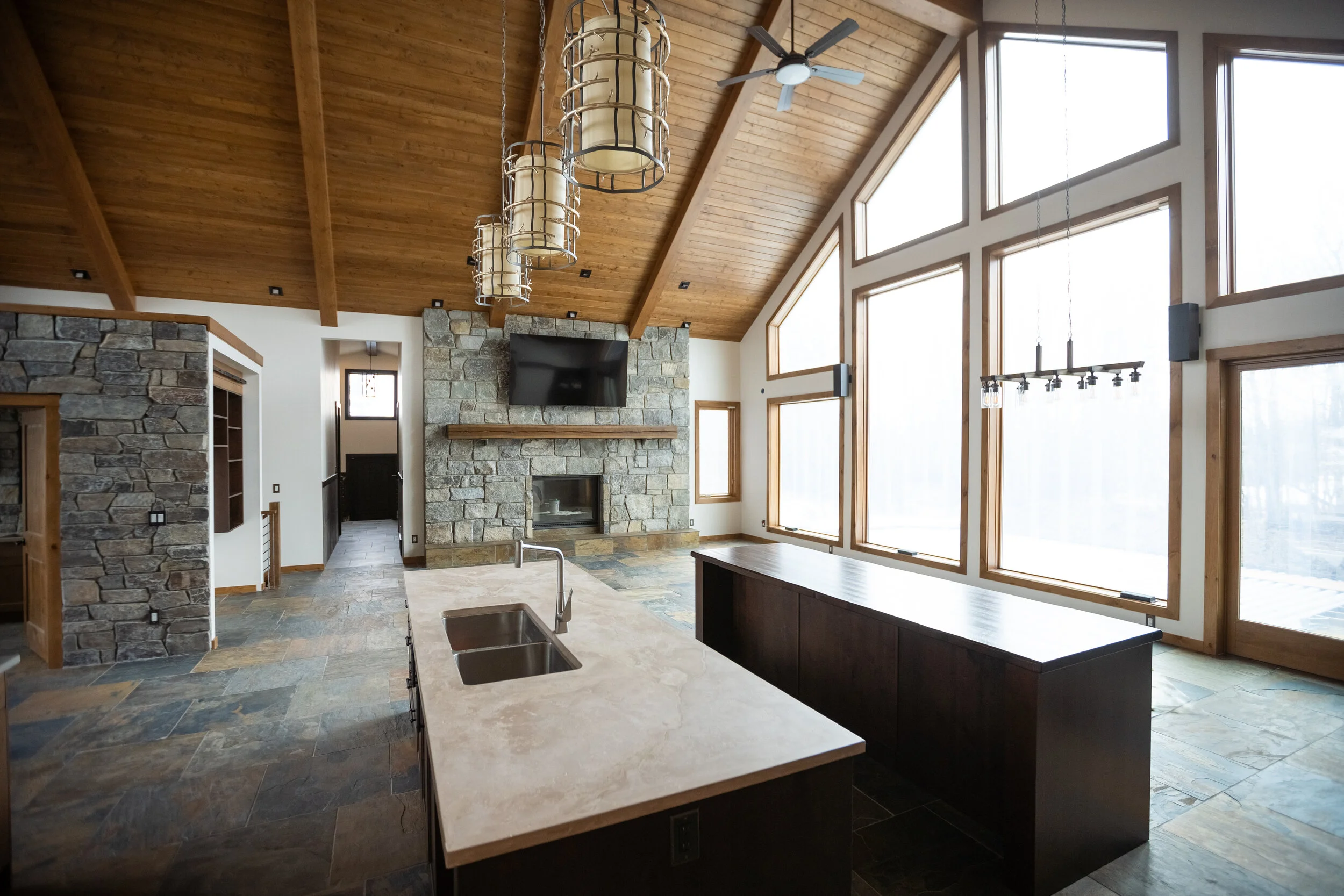 Interior of a modern house with a high wooden vaulted ceiling, large windows, a stone fireplace with a mounted TV above, and a kitchen island with a sink. Wood accents and contemporary lighting fixtures are visible.
