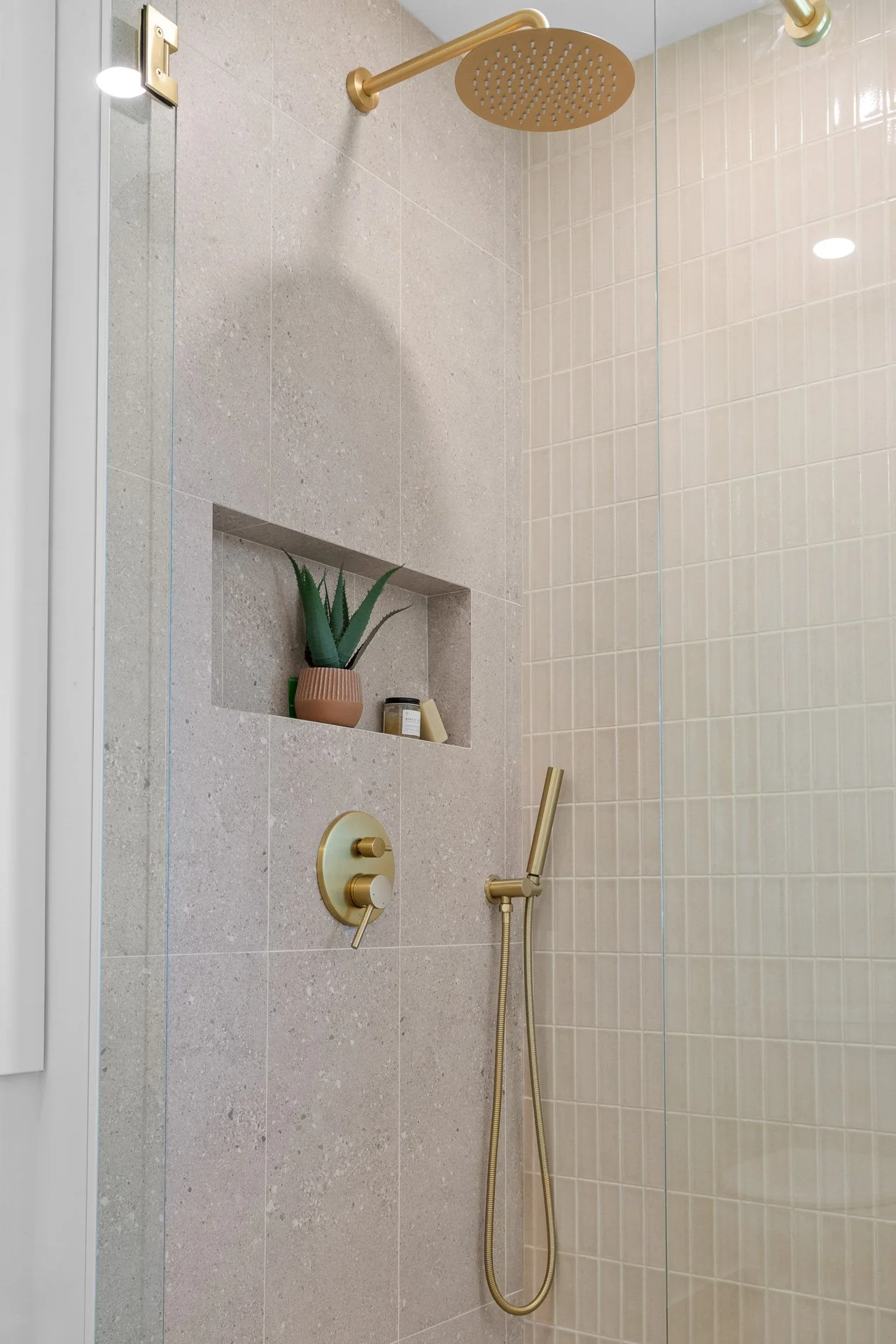 Shower with beige and cream tiles, gold fixtures, built-in shelf with a potted plant, soap, and lotion, glass shower door, and rain showerhead.