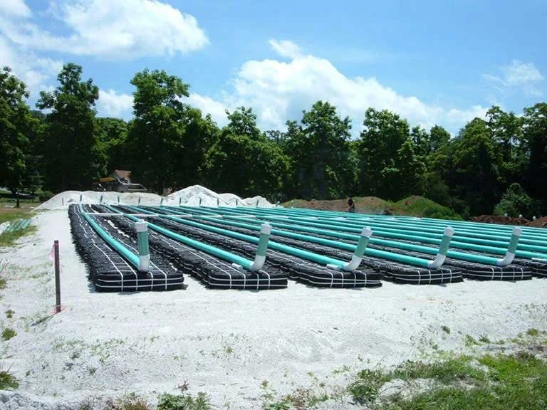 Installation of Eljen GSF septic system on a construction site, with black base mats and blue pipes laid out on gravel, surrounded by trees and blue skies.