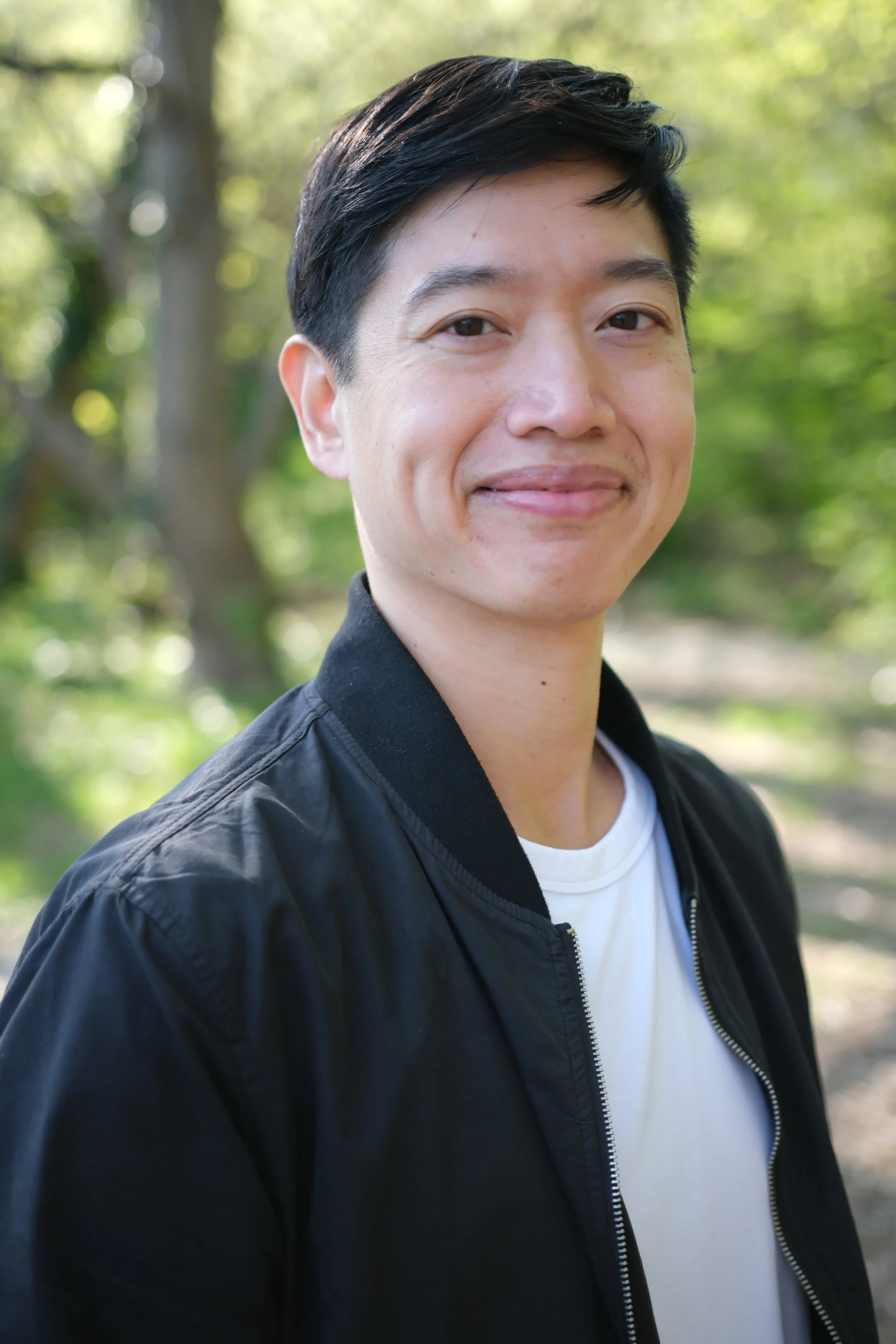 Portrait of a young man outdoors with sunlight filtering through trees in the background, wearing a black jacket and white shirt, smiling softly.