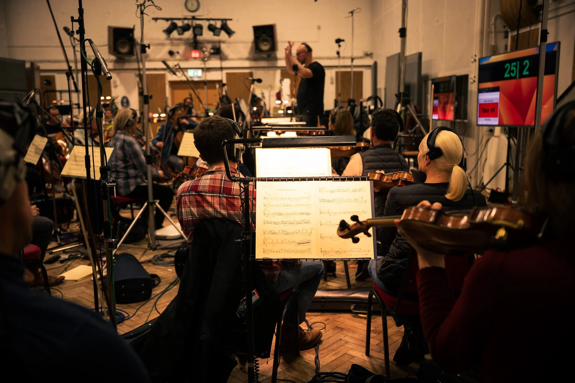 Orchestra rehearsal in recording studio with musicians playing violins and conductor on elevated platform.