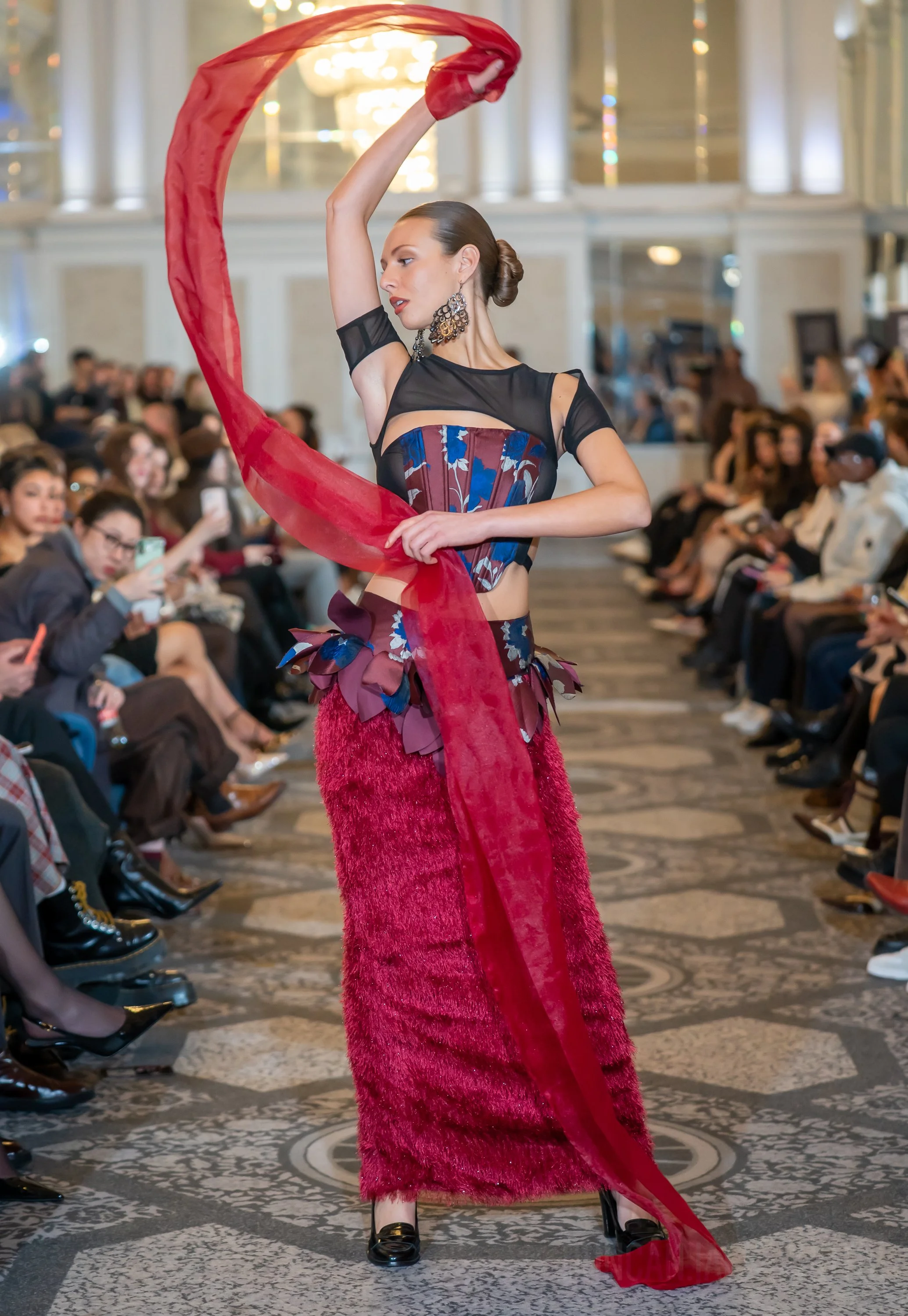 A woman wearing an elaborate red and blue fashion ensemble with a long red sheer scarf performing a dance or modeling on a runway in front of an audience.