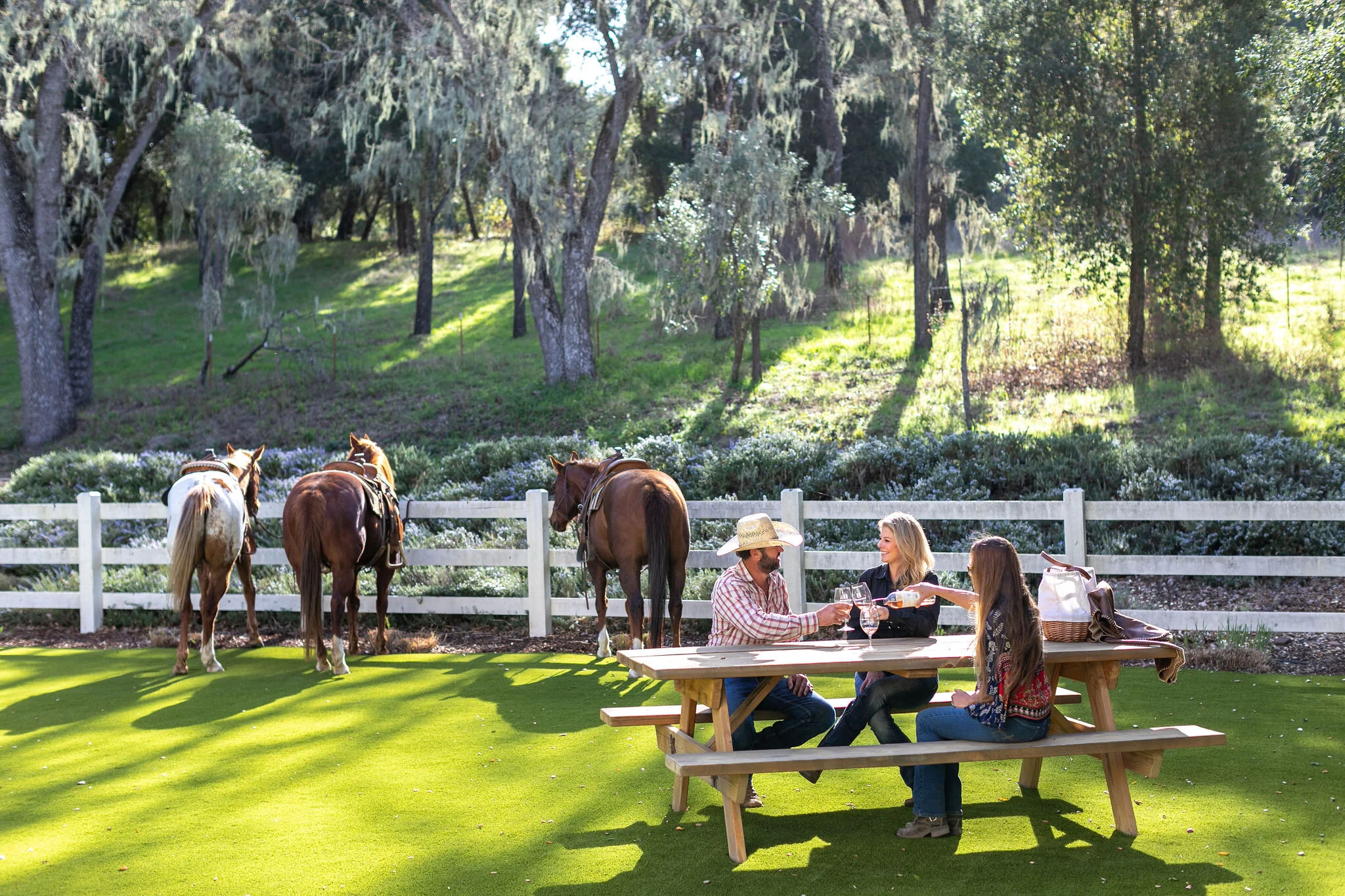 People sitting at a picnic table outdoors, with horses grazing behind a white fence, and a wooded area with trees in the background.
