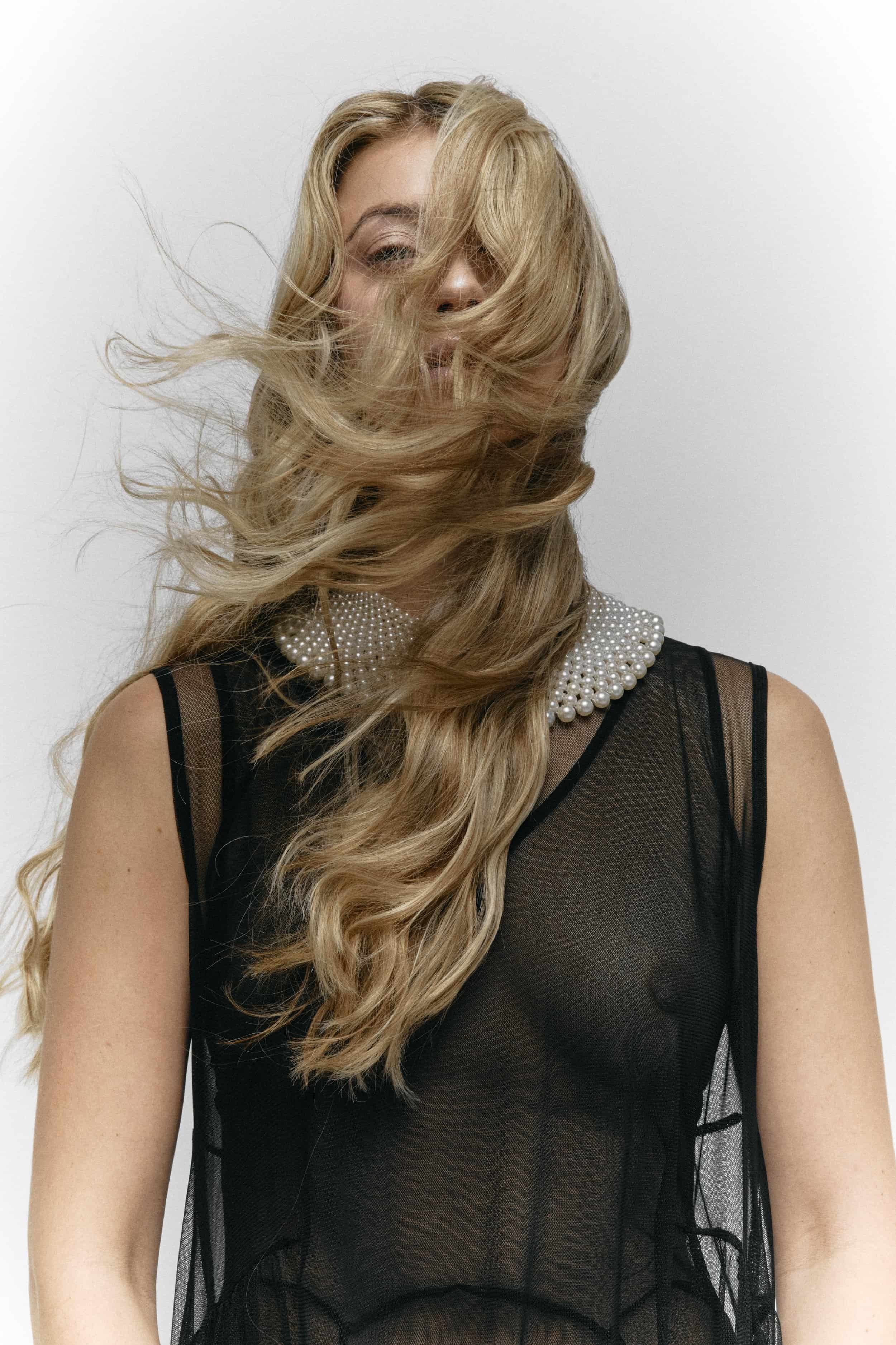 A woman with long, wavy blonde hair covering part of her face, wearing a black mesh top with a pearl necklace collar, standing against a plain white background.