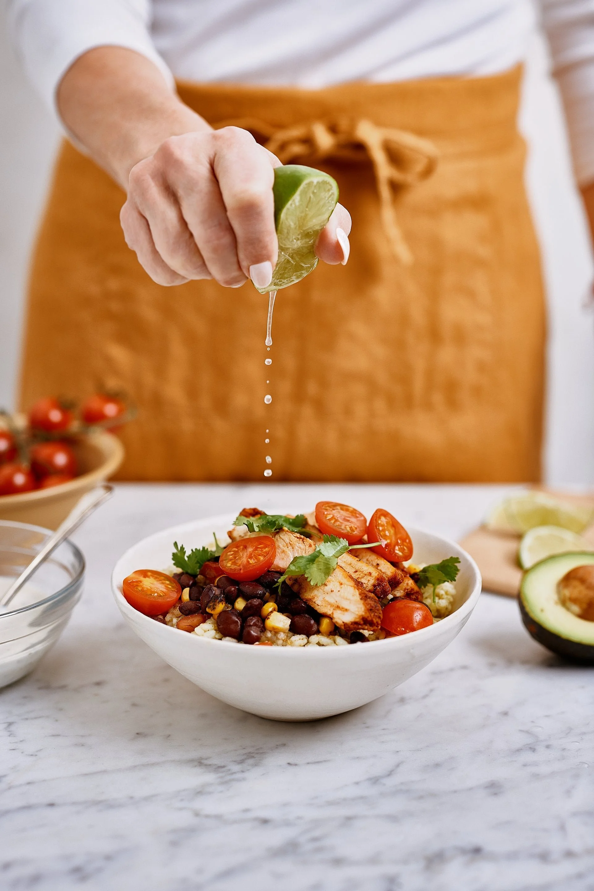 A bowl of salad topped with cherry tomatoes, grilled chicken, black beans, corn, and cilantro, with a person squeezing lime over it in a bright kitchen setting.