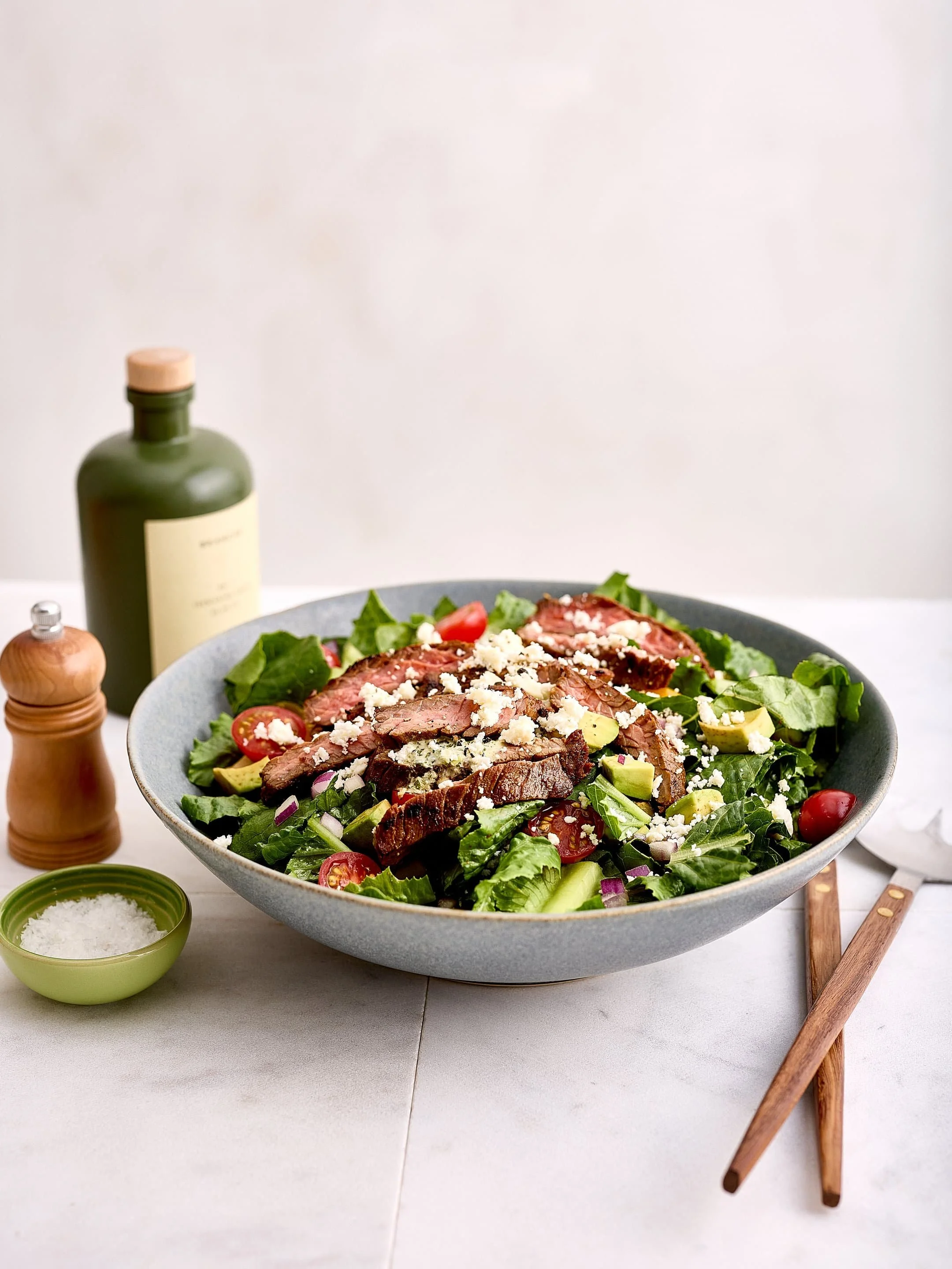 A large bowl of salad with lettuce, cherry tomatoes, avocado, red onions, grilled steak slices, and crumbled cheese. Nearby are bottles of salad dressing, salt, and pepper, on a white table with a wood-handled fork.