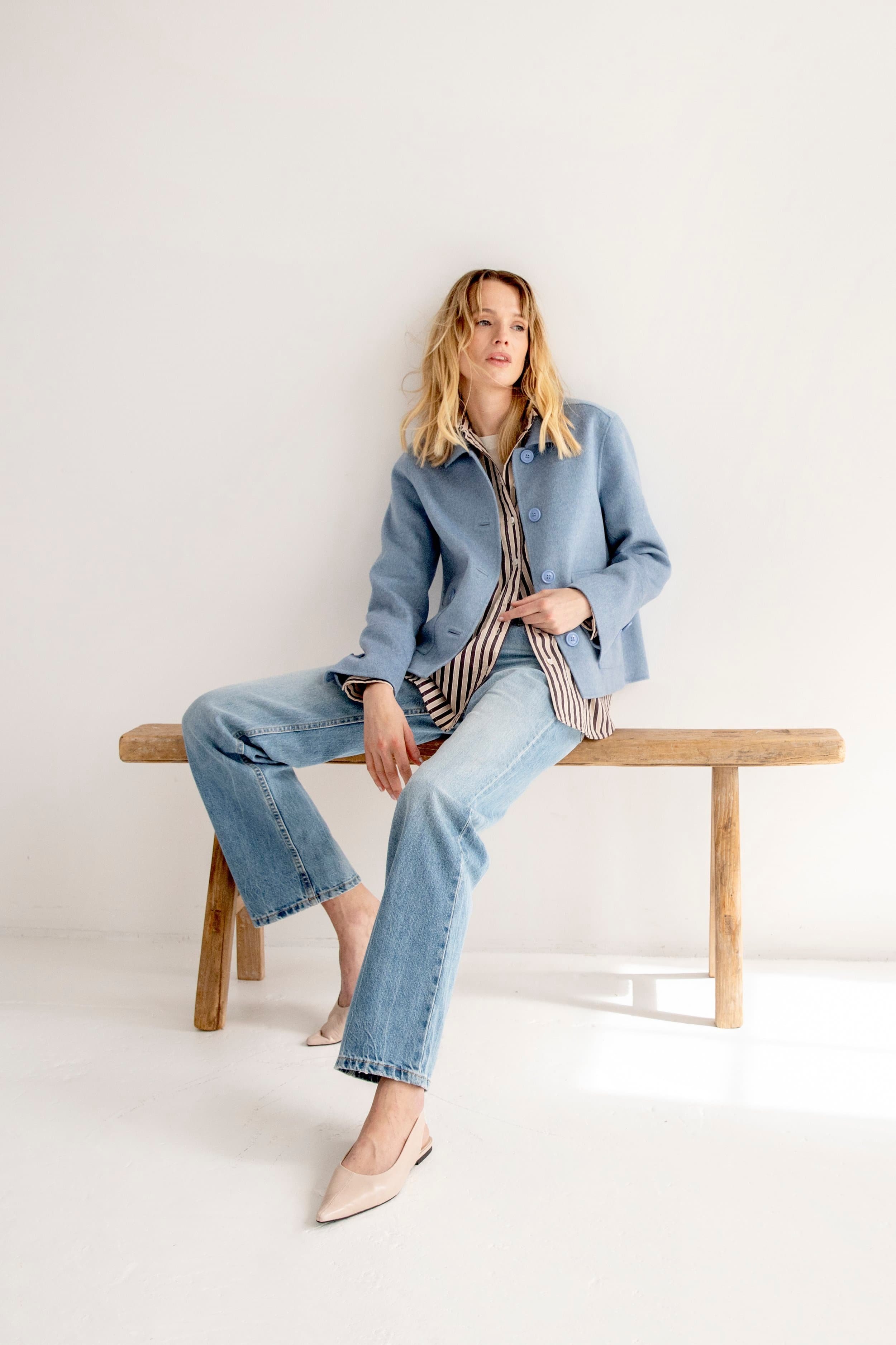 A woman with blonde hair wearing a blue jacket, striped shirt, and wide-leg jeans, sitting on a wooden bench in a minimal white room, with beige high heels.