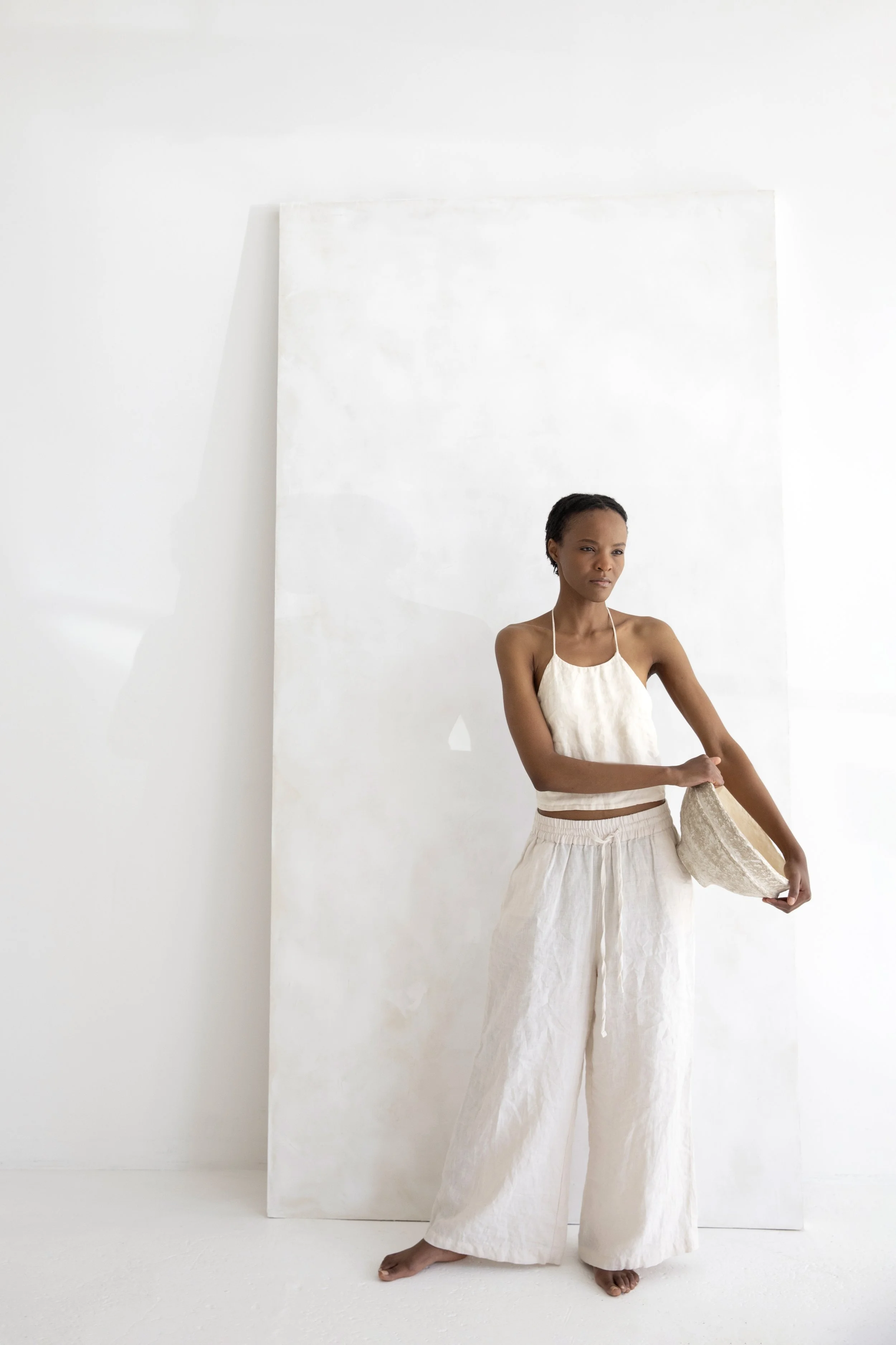 A woman with short hair wearing a beige halter top and wide-leg beige pants, holding a beige clutch, standing barefoot against a white background with a large white panel. Greater Boston Commercial Photo Studio